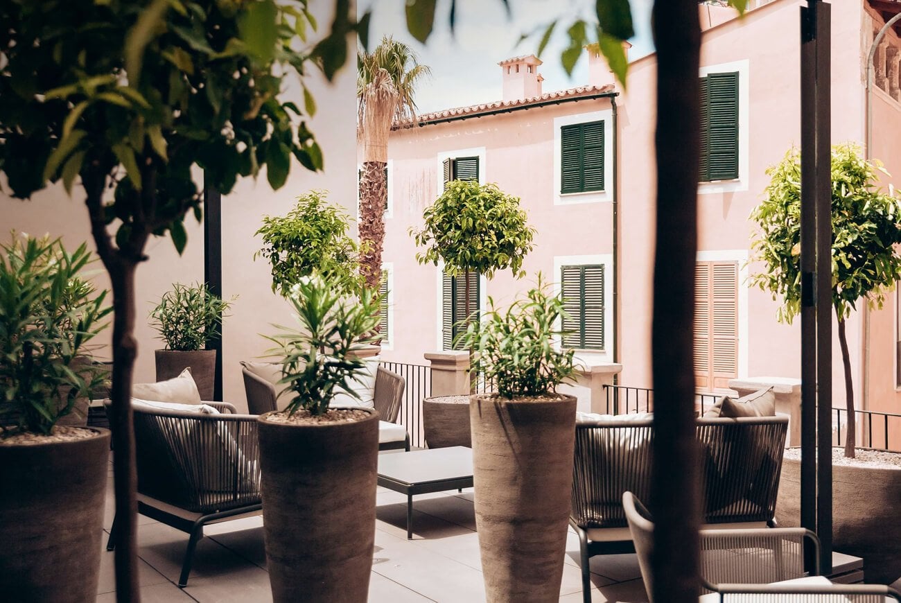Sant Francesc Hotel Singular, Balearic Islands, Spain — Courtyard with terracotta planters, woven chairs, palm trees, and pink-stucco Mediterranean architecture with green shutters.