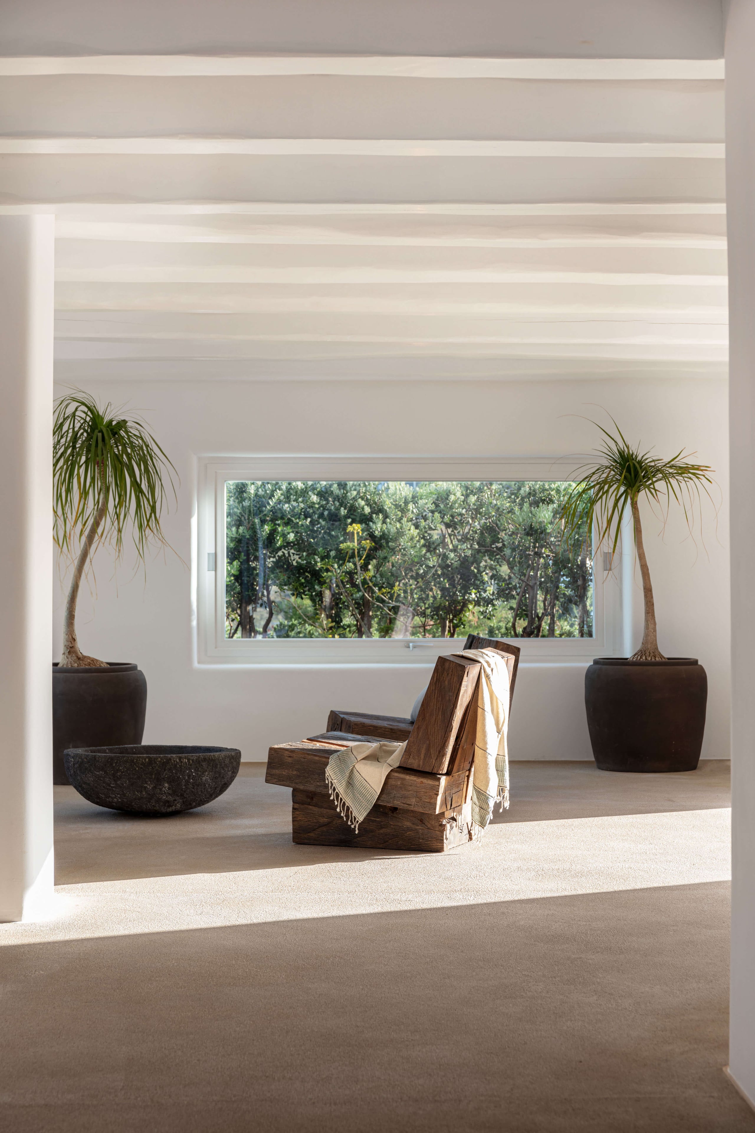 Villa Tama, Europe — Minimalist sunlit room with wooden lounge chair, potted palms, stone bowl, and expansive window framing forest views.