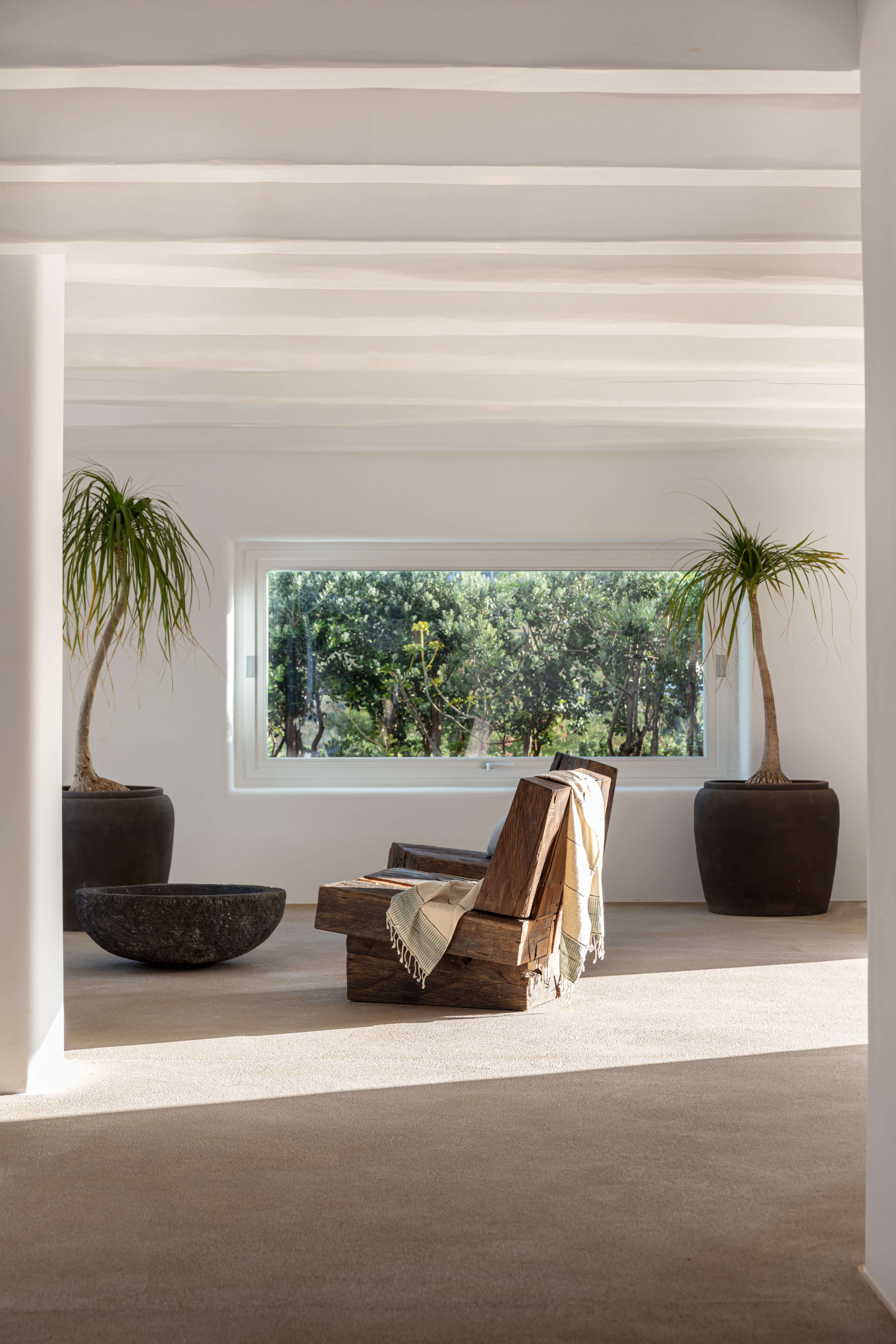 Villa Tama, Europe — Minimalist sunlit room with wooden lounge chair, potted palms, stone bowl, and expansive window framing forest views.