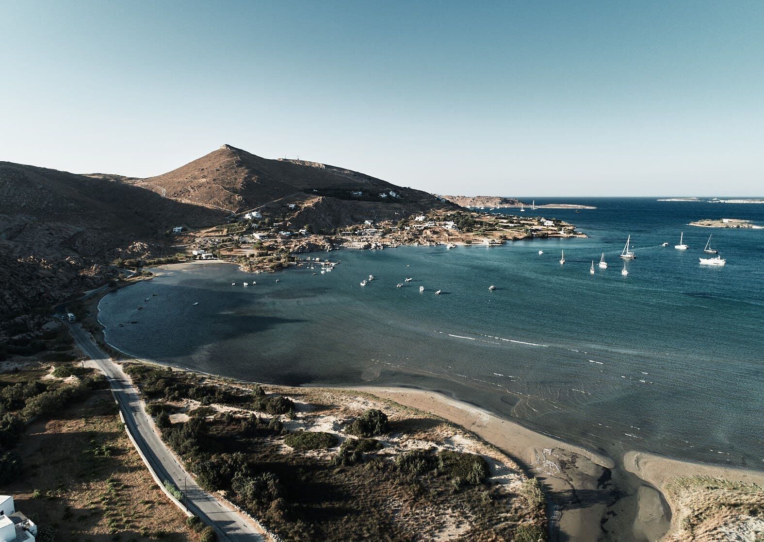 Parīlio Hotel, Europe — Coastal bay with moored sailboats, whitewashed village nestled against volcanic mountain, turquoise Mediterranean waters, golden afternoon light.