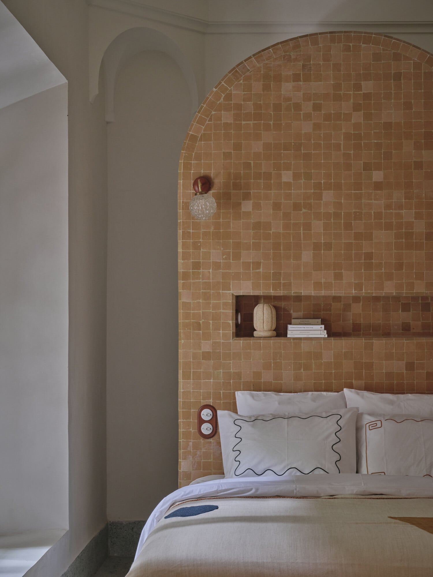 Rosemary, Africa — Arched terracotta tile headboard with recessed shelving above bed dressed in cream linens with wavy piping detail.