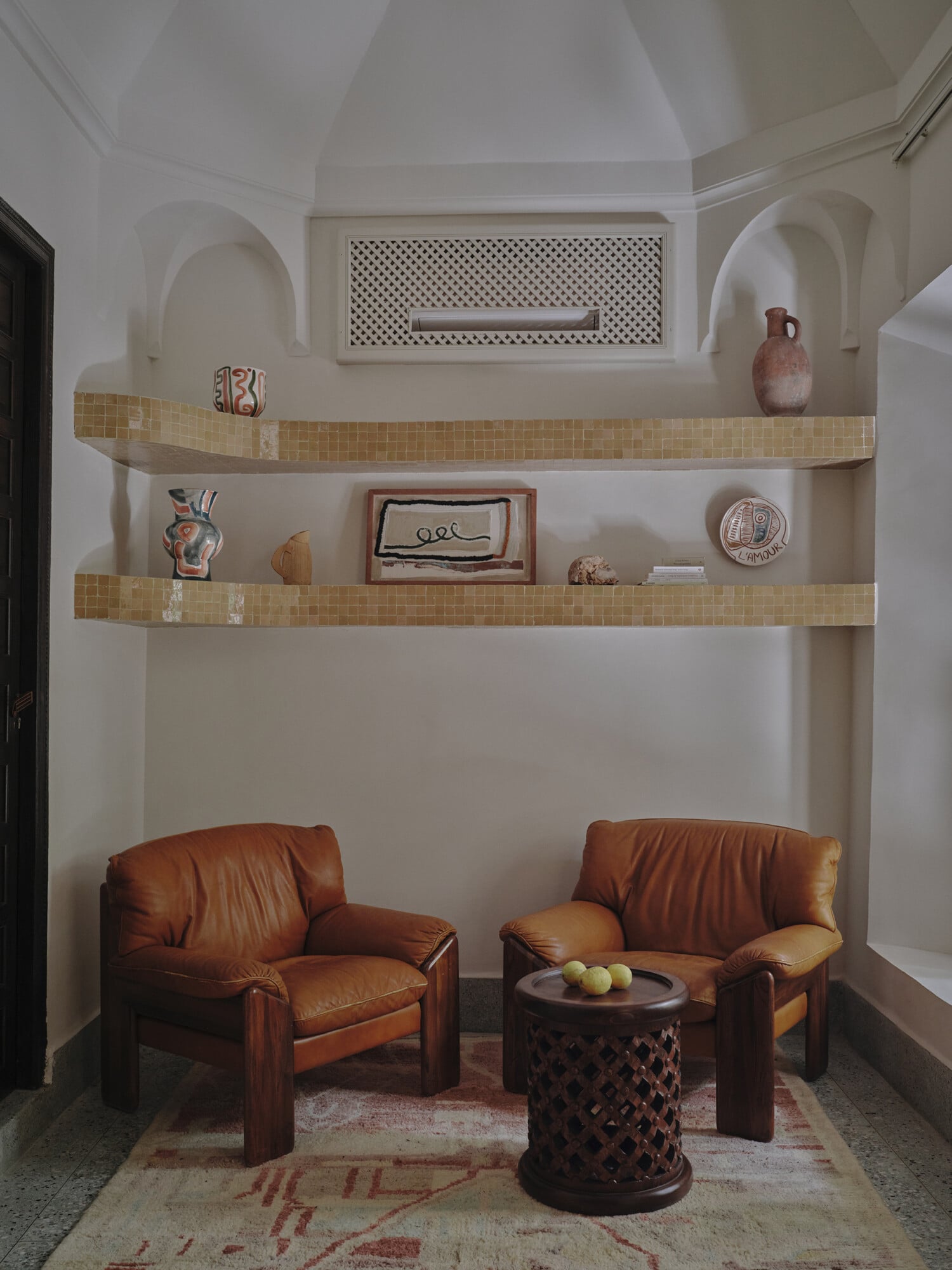 Rosemary, Africa — Sunlit nook with cognac leather chairs, mosaic tile shelves displaying ceramics, arched alcoves, carved wooden side table, minimalist hospitality design.