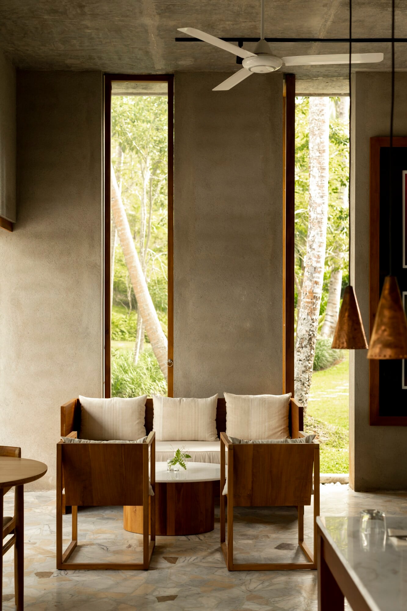 The Kitchen, Asia — Modernist lounge with walnut seating, cream cushions, concrete ceiling, pendant lights, and floor-to-ceiling windows framing tropical forest views.