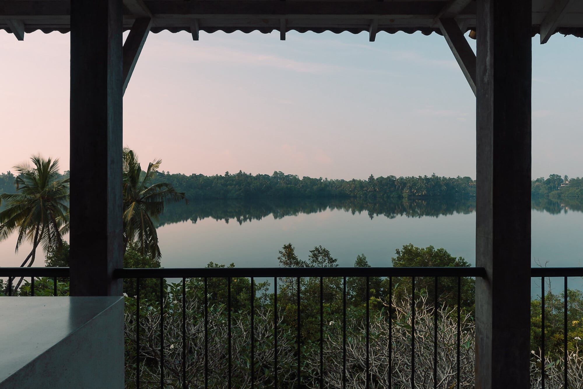 Kurulu Bay, Asia — Covered pavilion with metal railings overlooking a serene lake at dawn, tropical palms and forest beyond, calm water reflecting pale sky.
