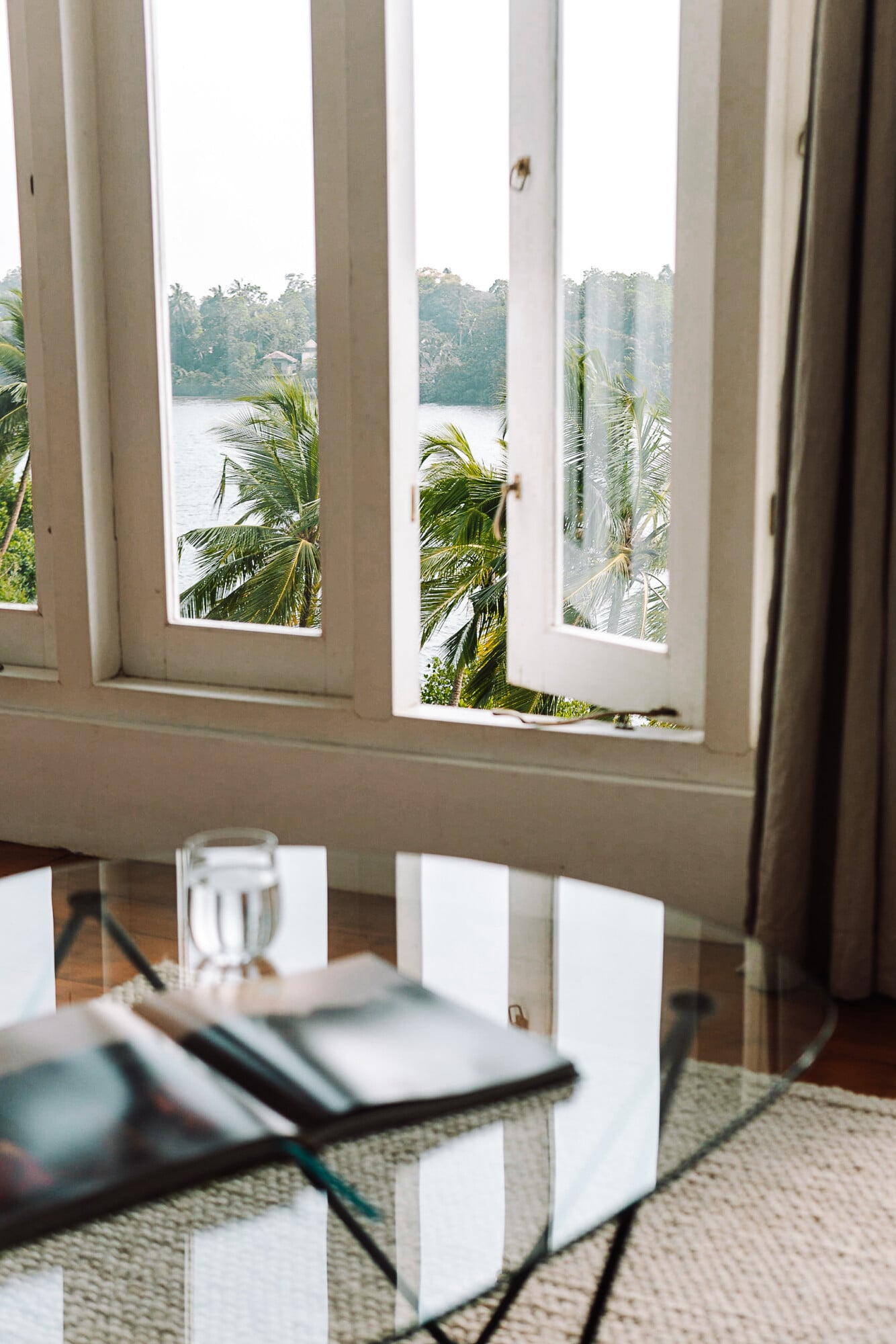 Kurulu Bay, Asia — Waterfront dining room with floor-to-ceiling windows framing palm trees and river views, natural light, minimalist white table setting, neutral linens and curtains.