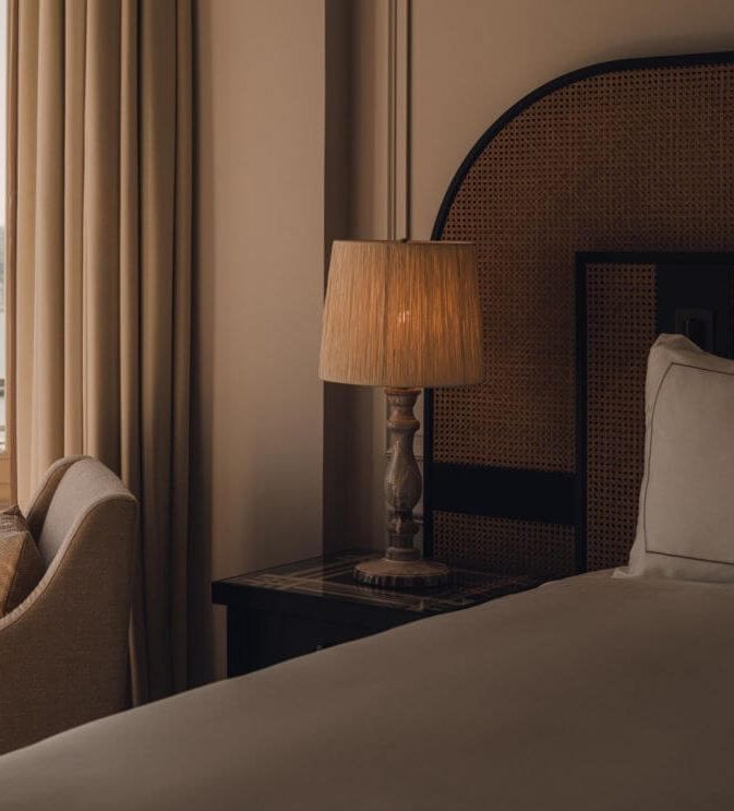 Carlton Cannes, Cannes, France — Luxury hotel bedroom with woven rattan headboard, wooden table lamp, beige linens, cream curtains, and warm ambient lighting.