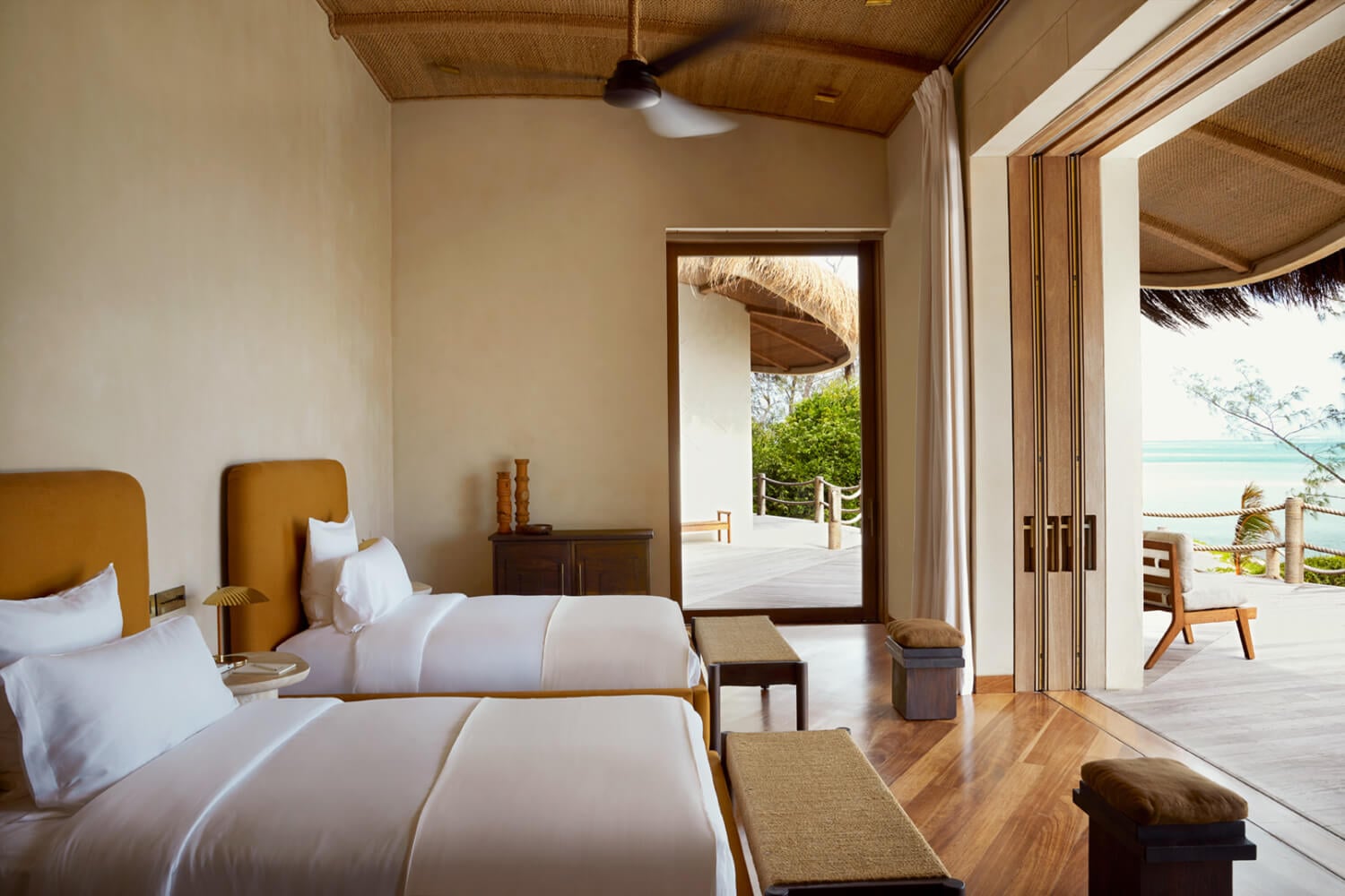 Kisawa Sanctuary, Africa — Twin beds with golden headboards, thatched ceiling, wooden floors, and ocean views through open doors to beachfront terrace.
