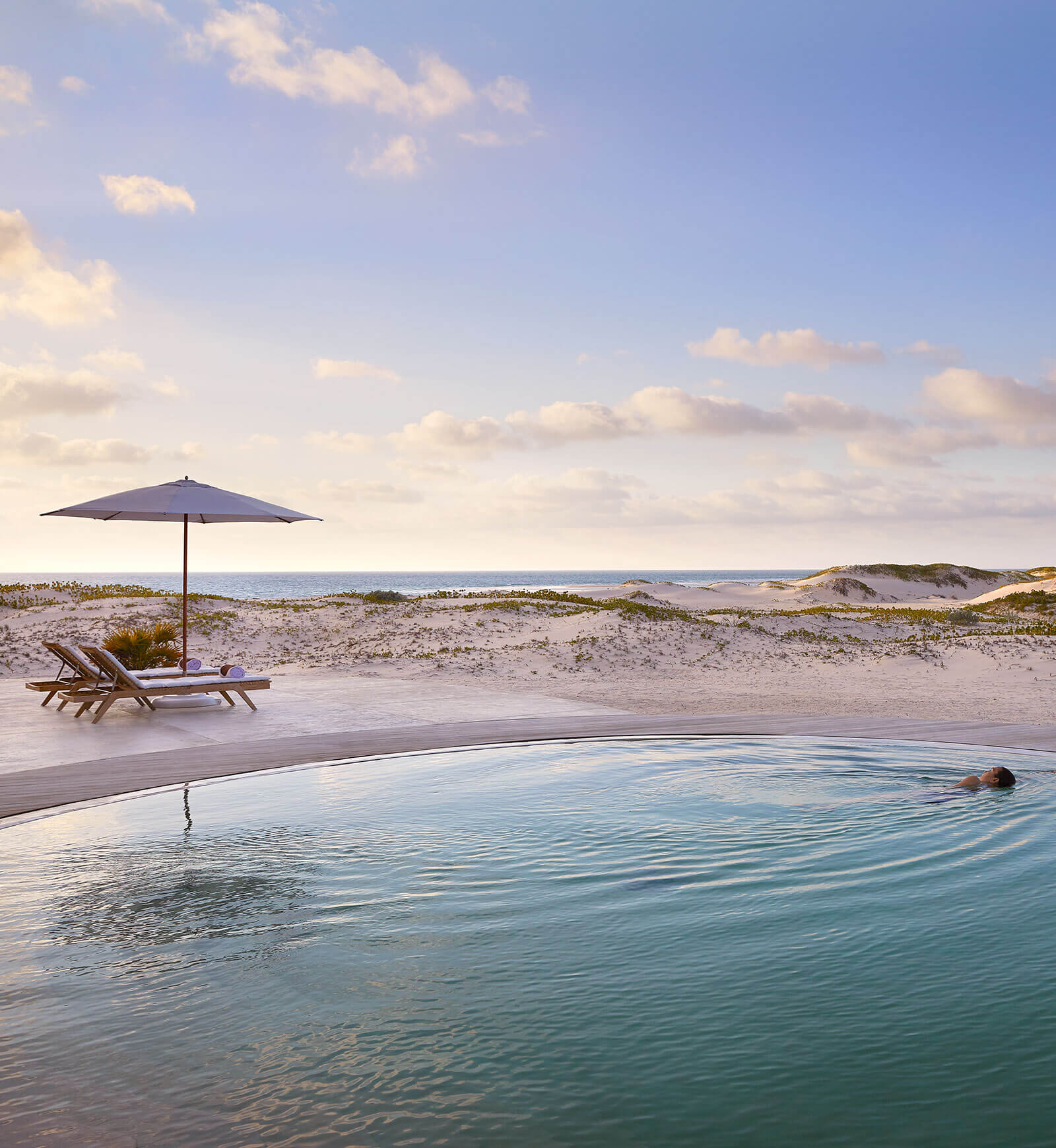 Lagoon Pool, Africa — infinity pool overlooking pristine beach with wooden loungers, blue umbrella, and sand dunes at golden hour.