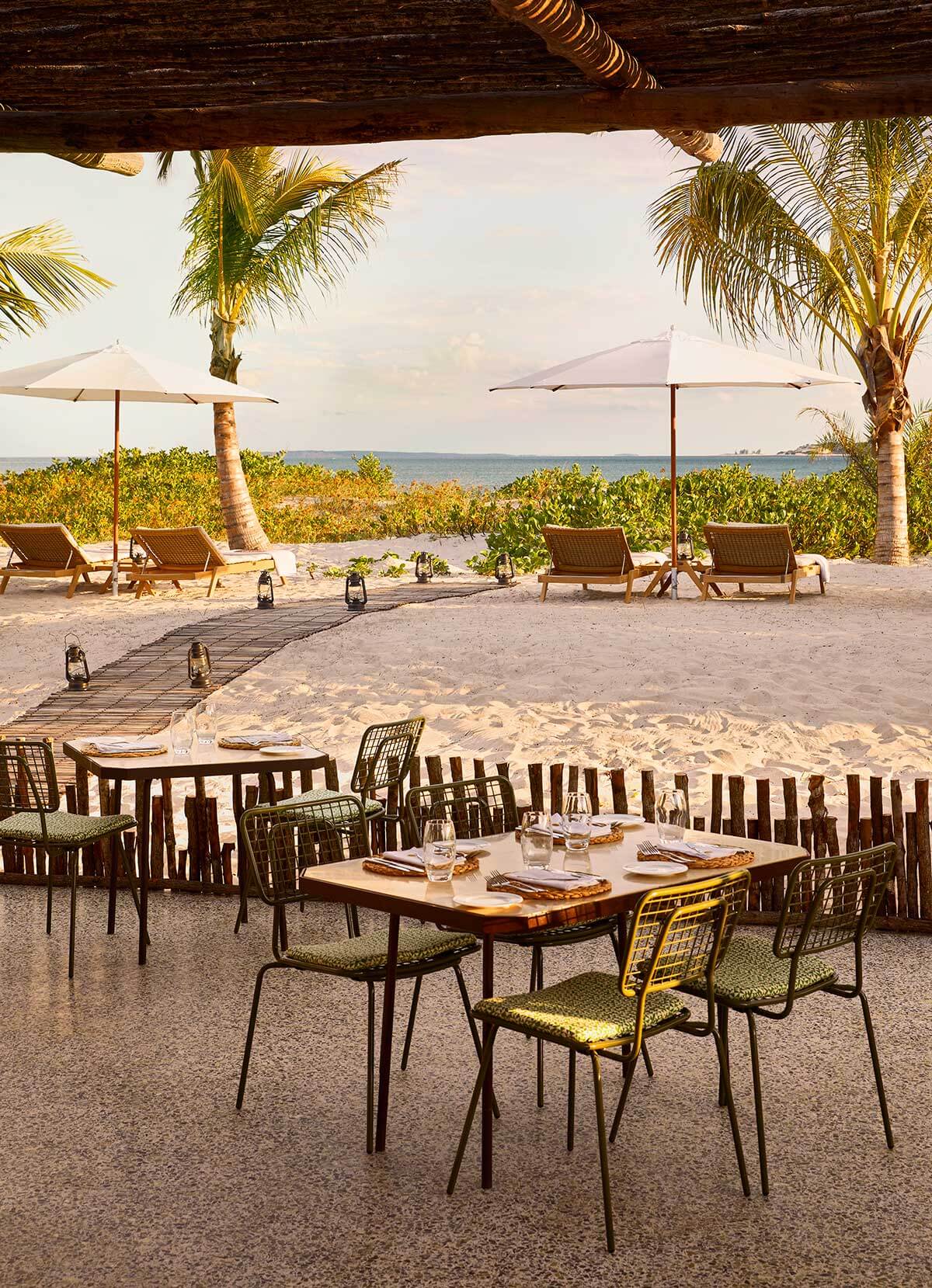 Ocean Mussassa, Africa — Beachfront dining pavilion with wooden table, modern chairs, and umbrellas overlooking ocean, palm trees, and mangrove vegetation at sunset.
