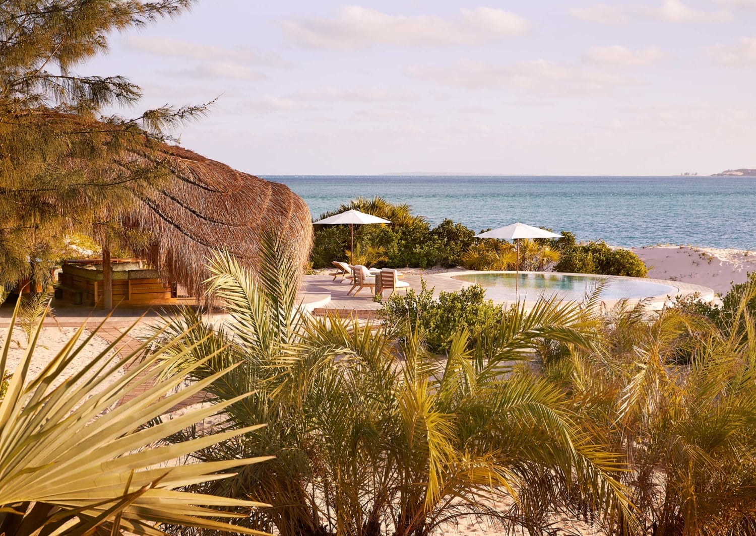 Kisawa Sanctuary, Africa — Thatched beach pavilion with infinity pool overlooking turquoise ocean, palm fronds, white umbrellas, wooden decking, tropical luxury resort architecture.