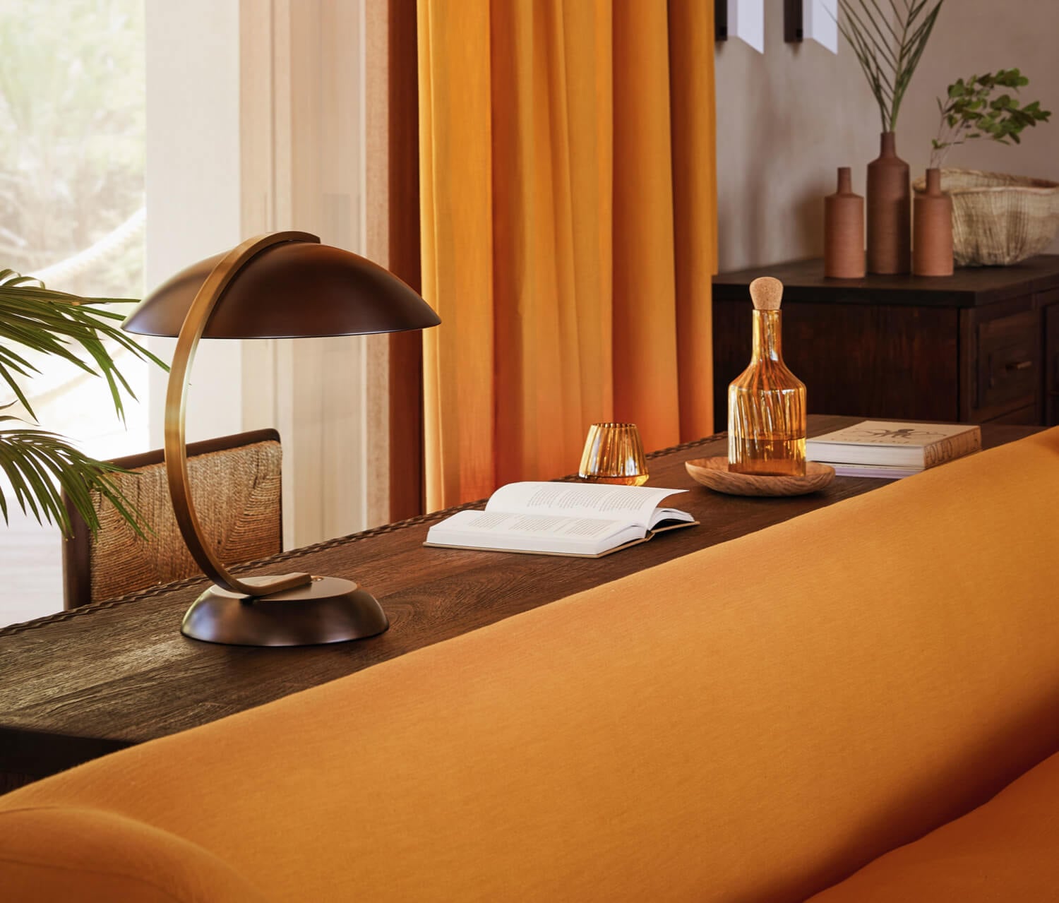 Kisawa Sanctuary, Africa — Warm-toned workspace with curved brass desk lamp, open book, amber glass decanter, and golden curtains filtering natural light over dark wood surface.