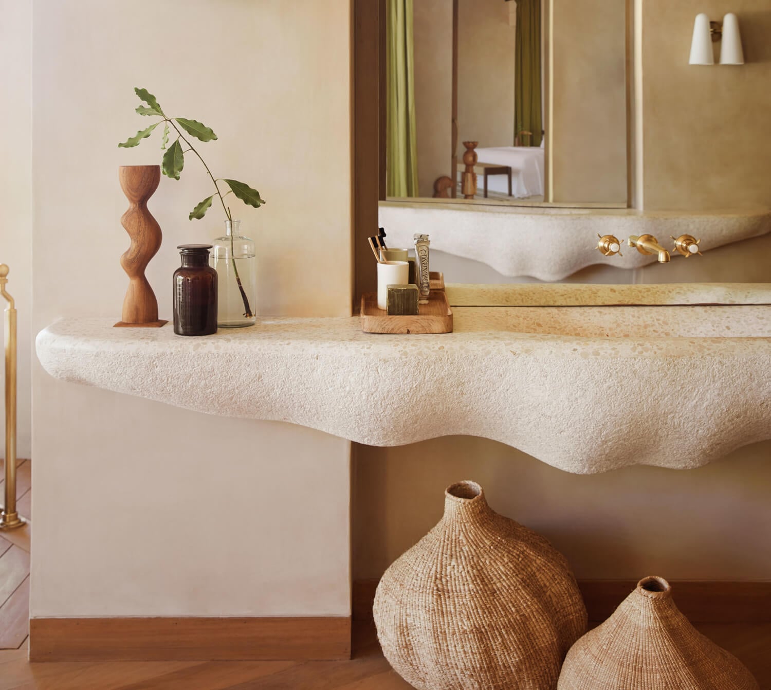 Kisawa Sanctuary, Africa — Curved cream limestone sink with wavy edge, wooden sculptural vessels, dark glass bottles, brass fixtures, and woven baskets in serene spa bathroom.
