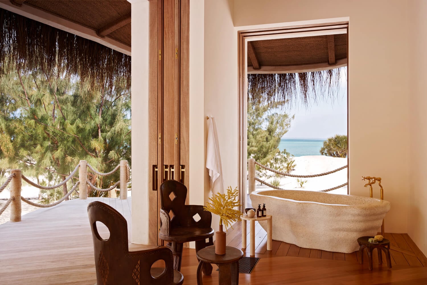 Kisawa Sanctuary, Africa — Luxury beach bathroom with sculptural wooden furniture, stone bathtub, and ocean views through floor-to-ceiling doors overlooking tropical landscape.