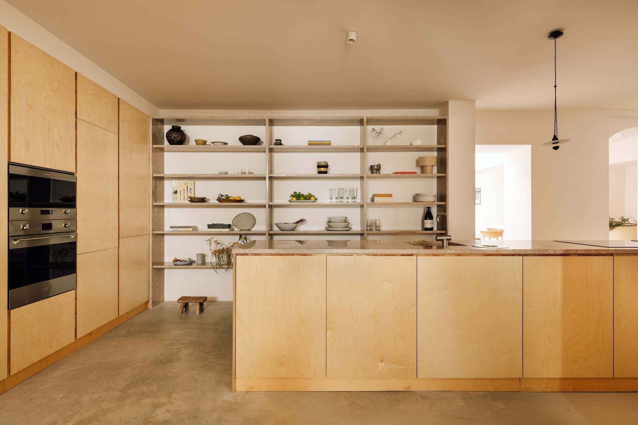 Casa Quatro, Europe — Minimalist kitchen with natural oak cabinetry, open shelving, stainless steel appliances, concrete flooring, and warm ambient light.