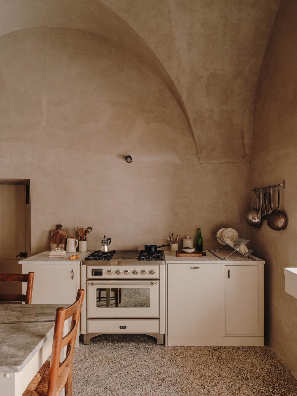 Casa Soleto, Europe — Minimalist kitchen with cream cabinetry, vintage range cooker, terracotta walls, and arched ceiling detail in rustic Italian villa.