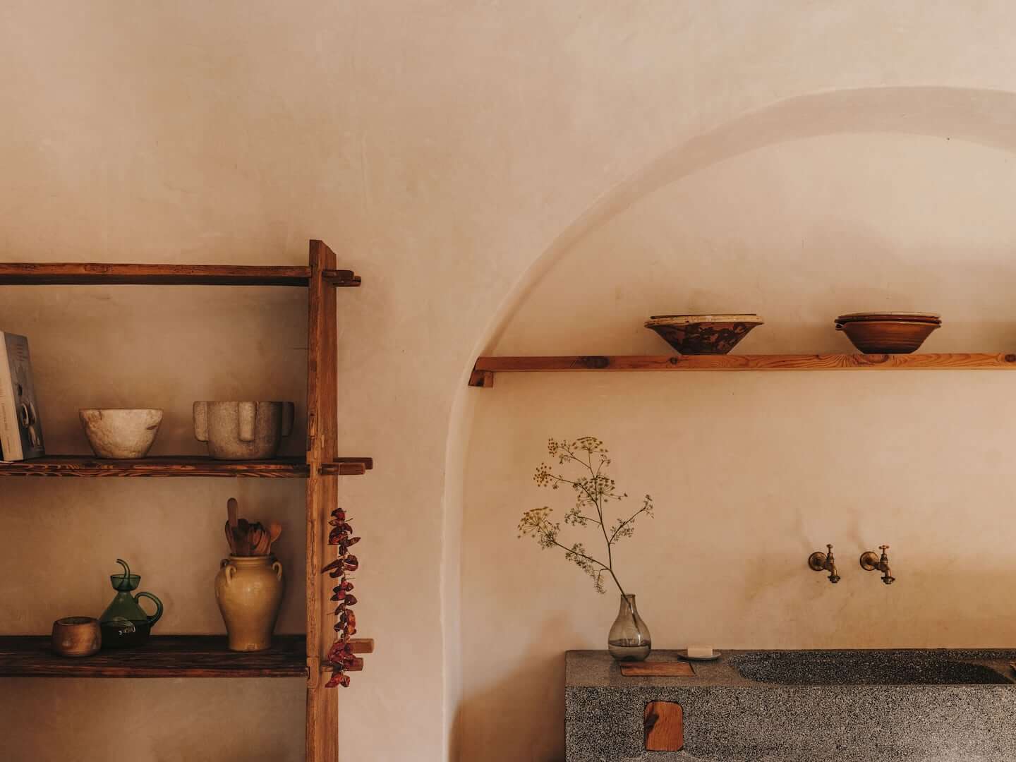 Can Lluïssó, Balearic Islands, Spain — Terracotta walls with wooden shelving displaying ceramics, dried botanicals in glass vase, bronze faucets, arched ceiling detail.