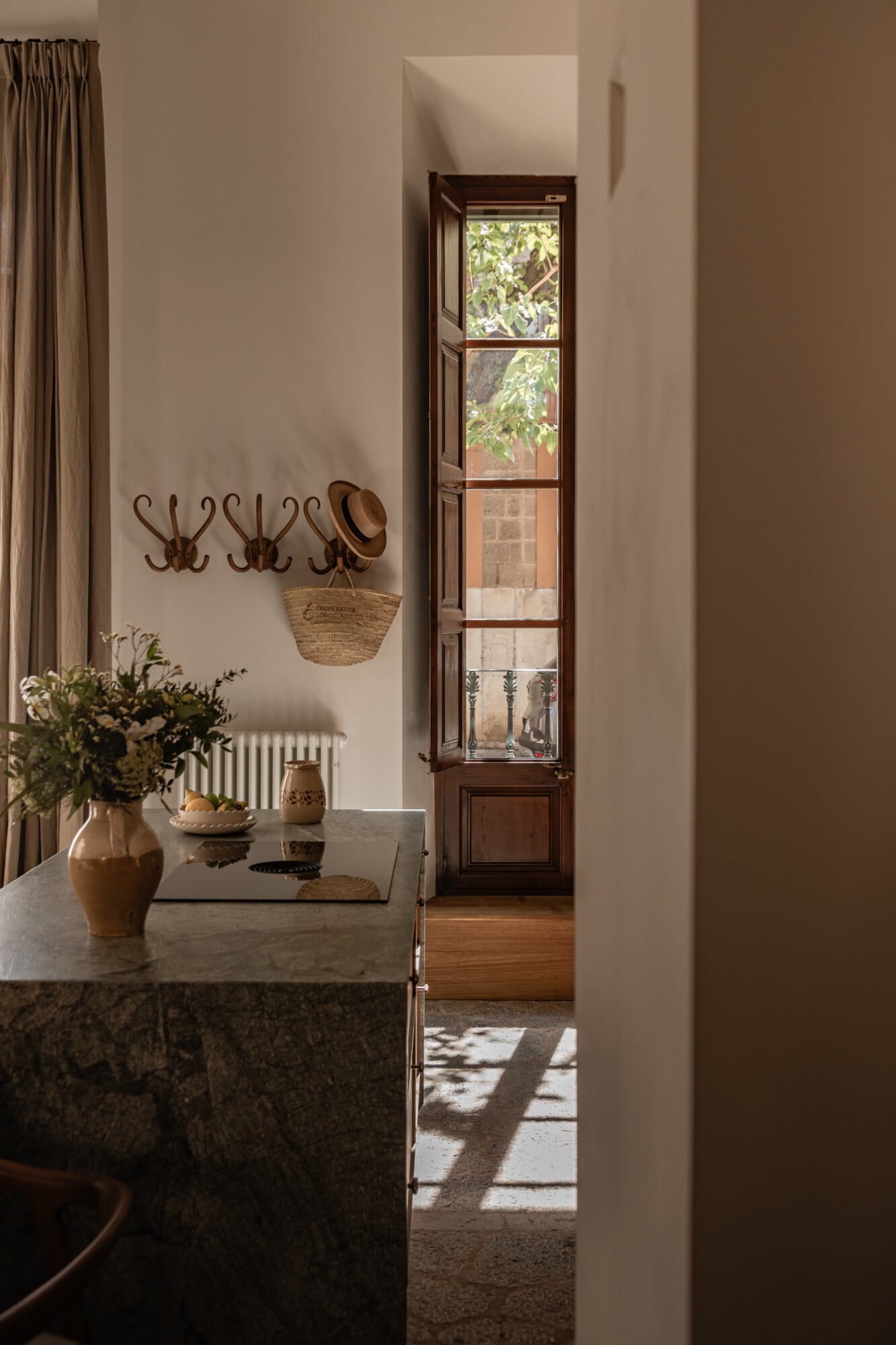 Mon Cor, Balearic Islands, Spain — Minimalist entryway with marble counter, wooden door, wall hooks, woven basket, potted flowers, and dappled sunlight through tall window.