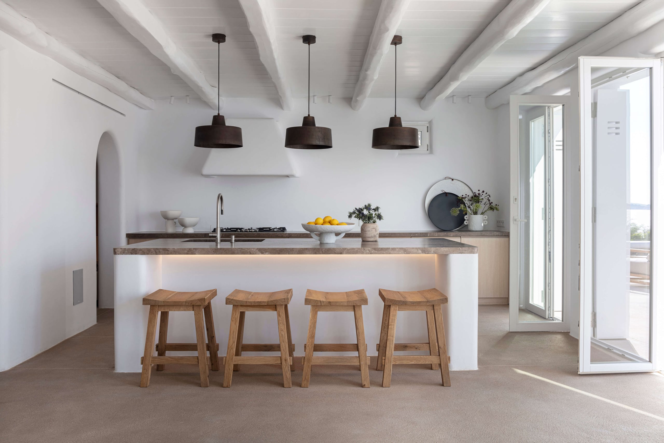 Villa Tama, Europe — Minimalist kitchen with white cabinetry, gray stone countertop, three black pendant lights, wooden bar stools, exposed ceiling beams, and glass doors opening to terrace.