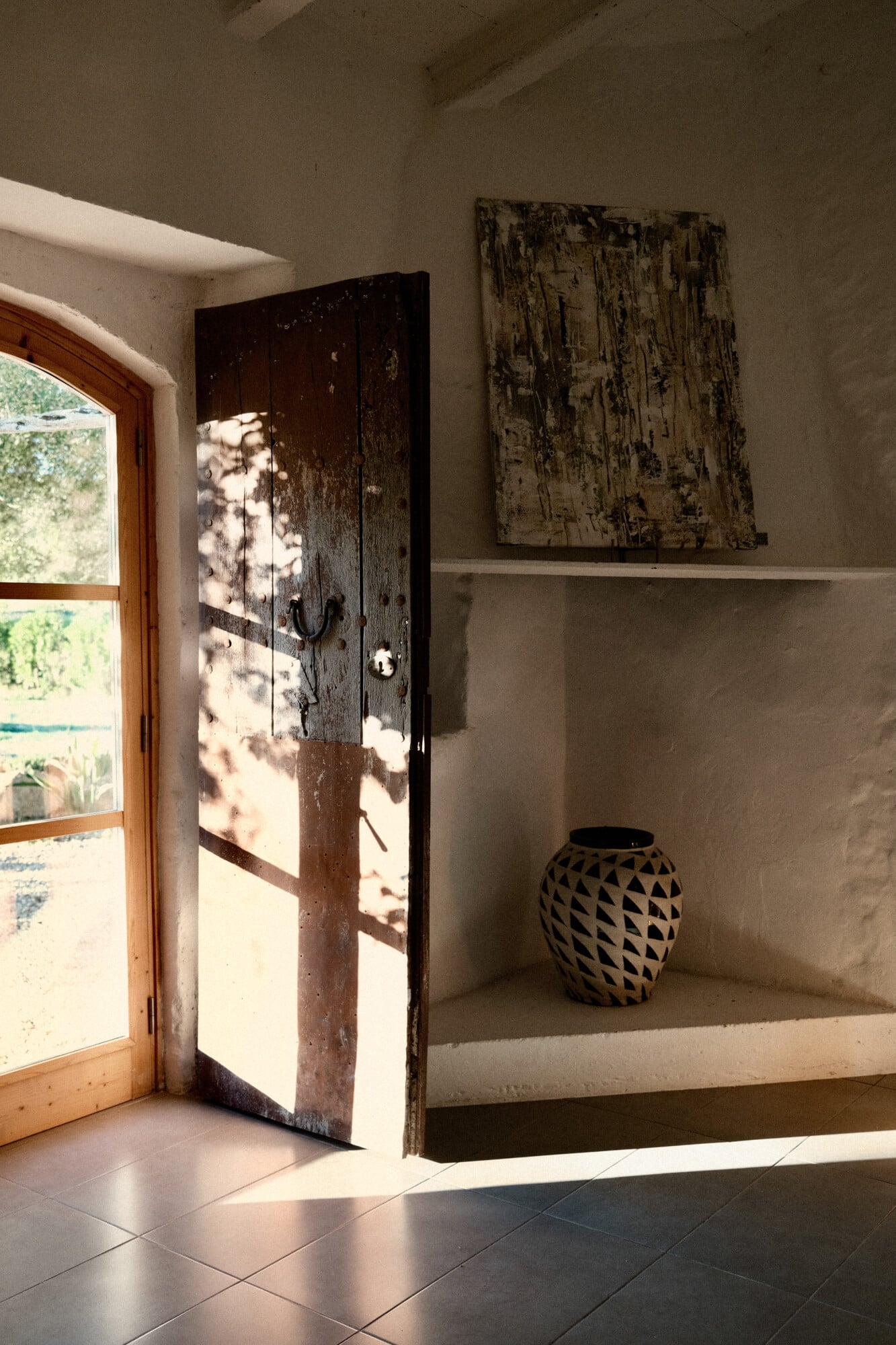Vista Verde, Balearic Islands — Weathered wooden door with dappled sunlight, artisan pottery vessel, and textured wall art in minimalist Mediterranean interior.
