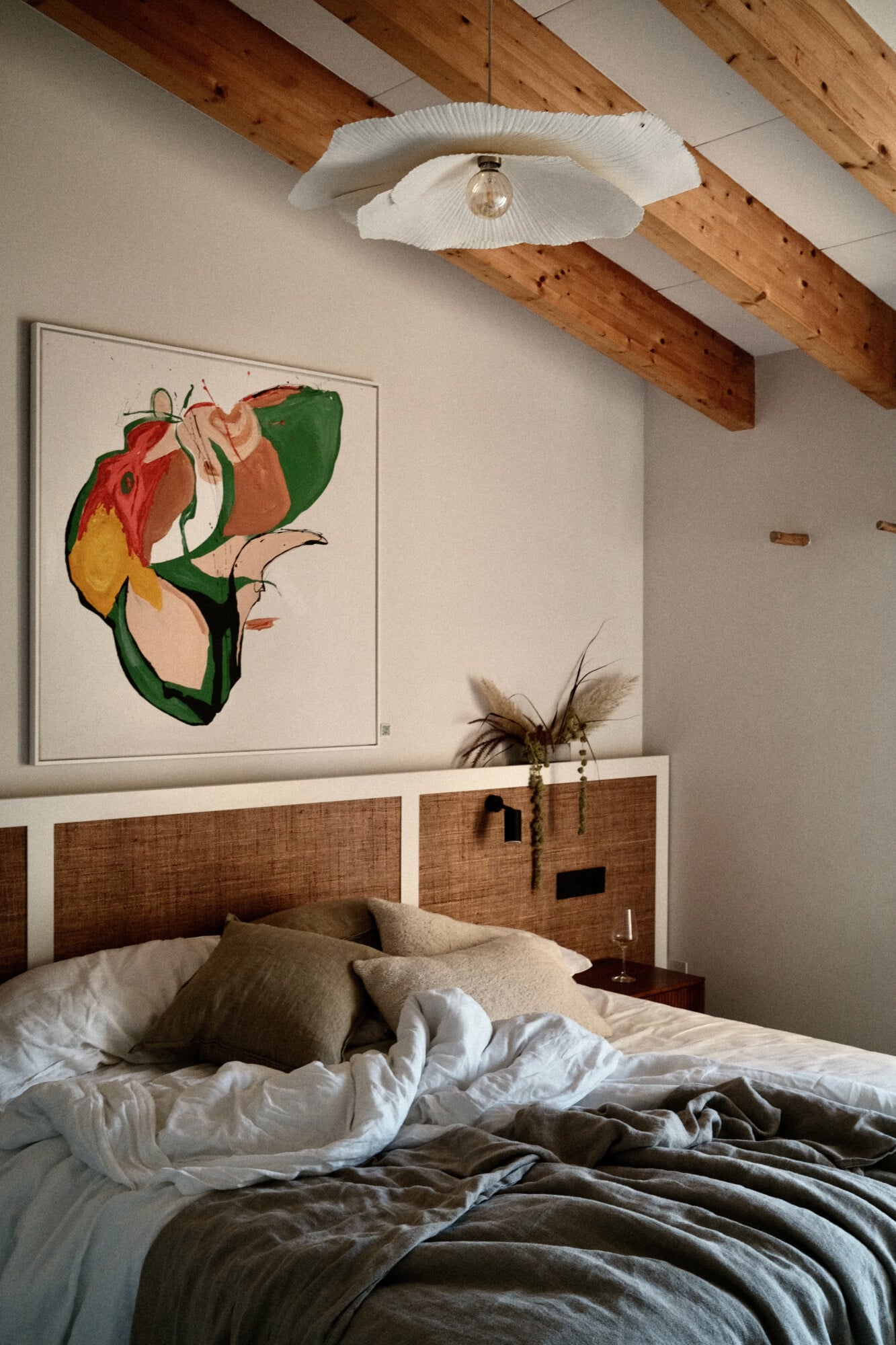 Vista Verde, Balearic Islands — Minimalist bedroom with woven rattan headboard, exposed wooden beams, botanical artwork, linen bedding, and sculptural pendant light.