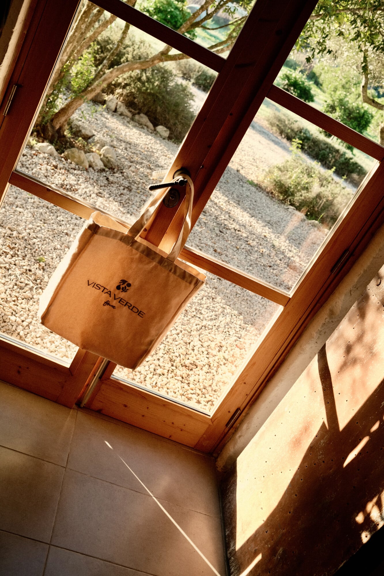 Vista Verde, Balearic Islands — Canvas tote bag hanging from wooden skylight frame overlooking gravel courtyard and Mediterranean vegetation, warm natural light.