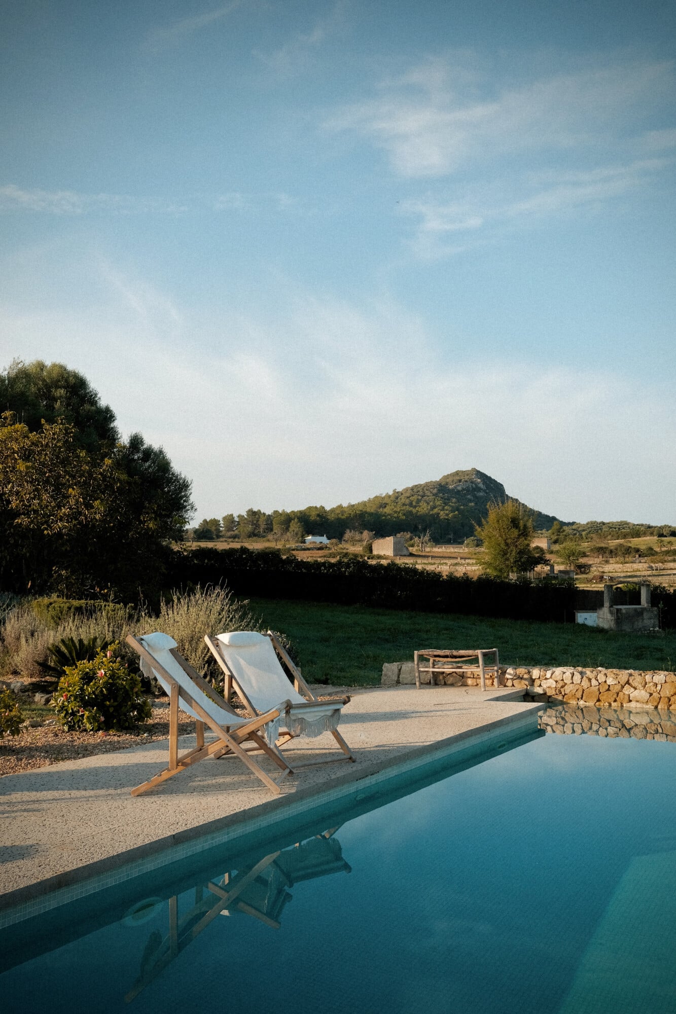 Vista Verde, Balearic Islands — Two linen deck chairs beside turquoise pool overlooking countryside, stone terrace, Mediterranean landscape with hills.