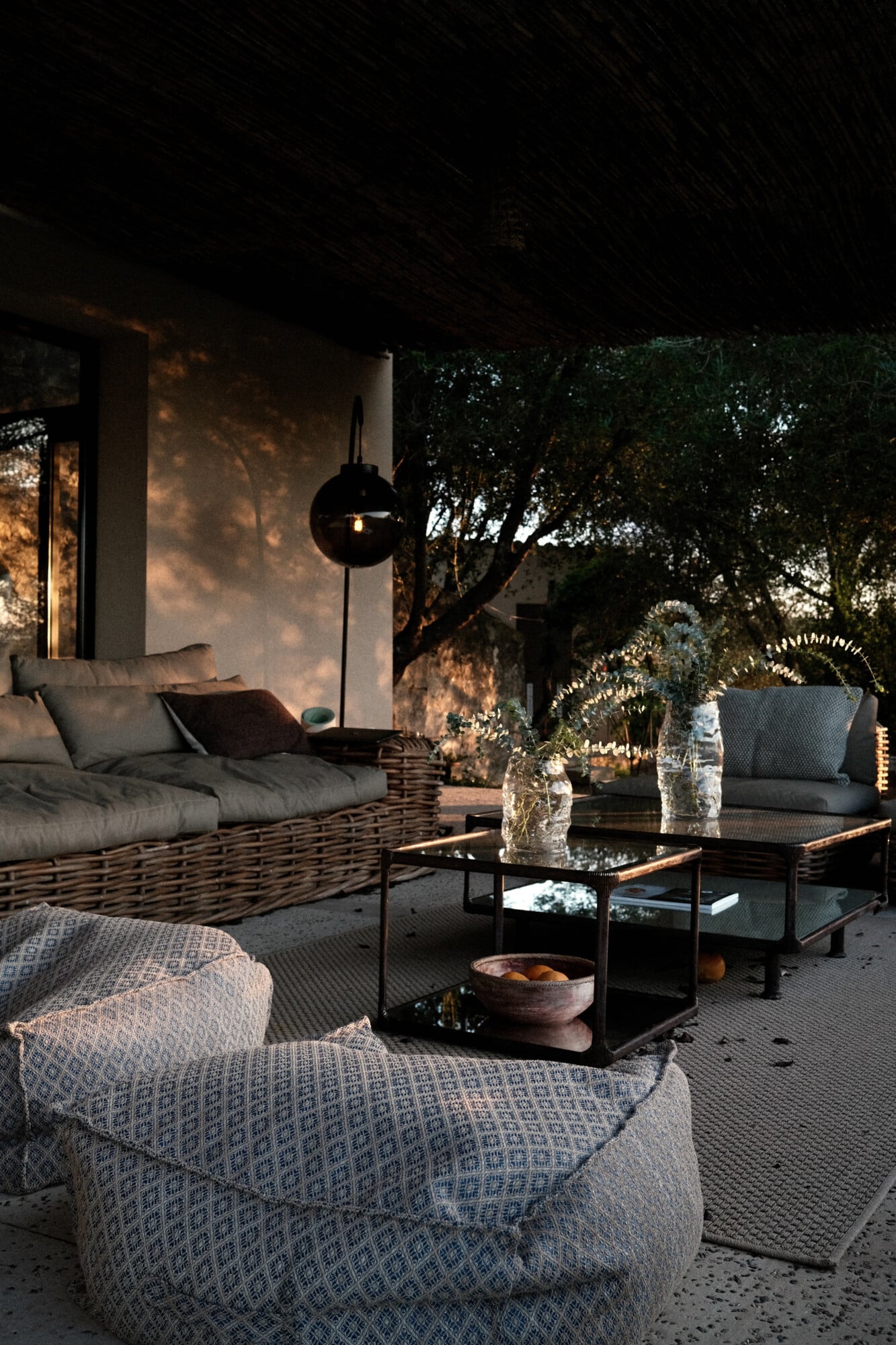 Vista Verde, Balearic Islands — Evening patio with woven lounge seating, black arc lamp, stone terrace, and olive trees casting dappled shadows.