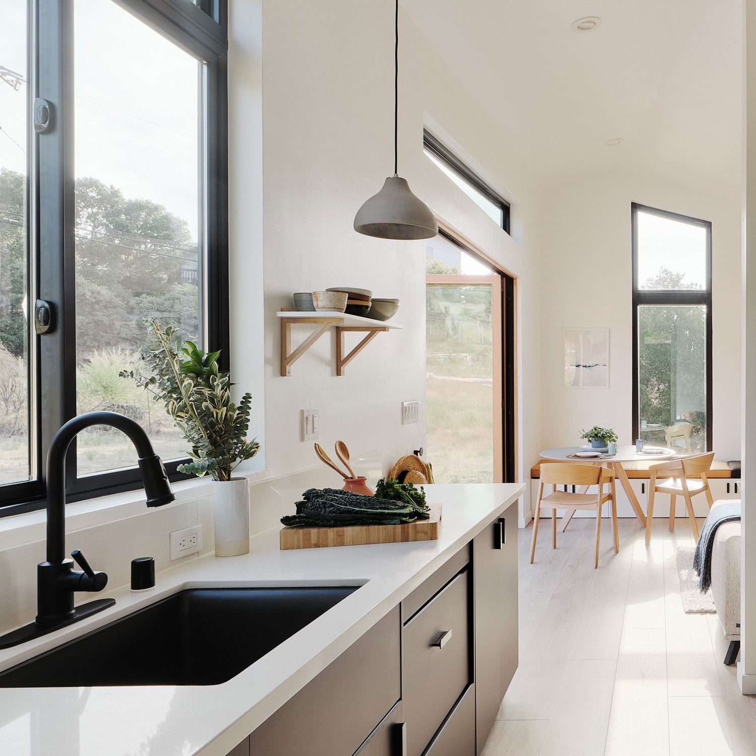 Abodu x Koto, North America — Minimalist kitchen with black sink, pendant light, and dining area overlooking countryside through floor-to-ceiling windows.