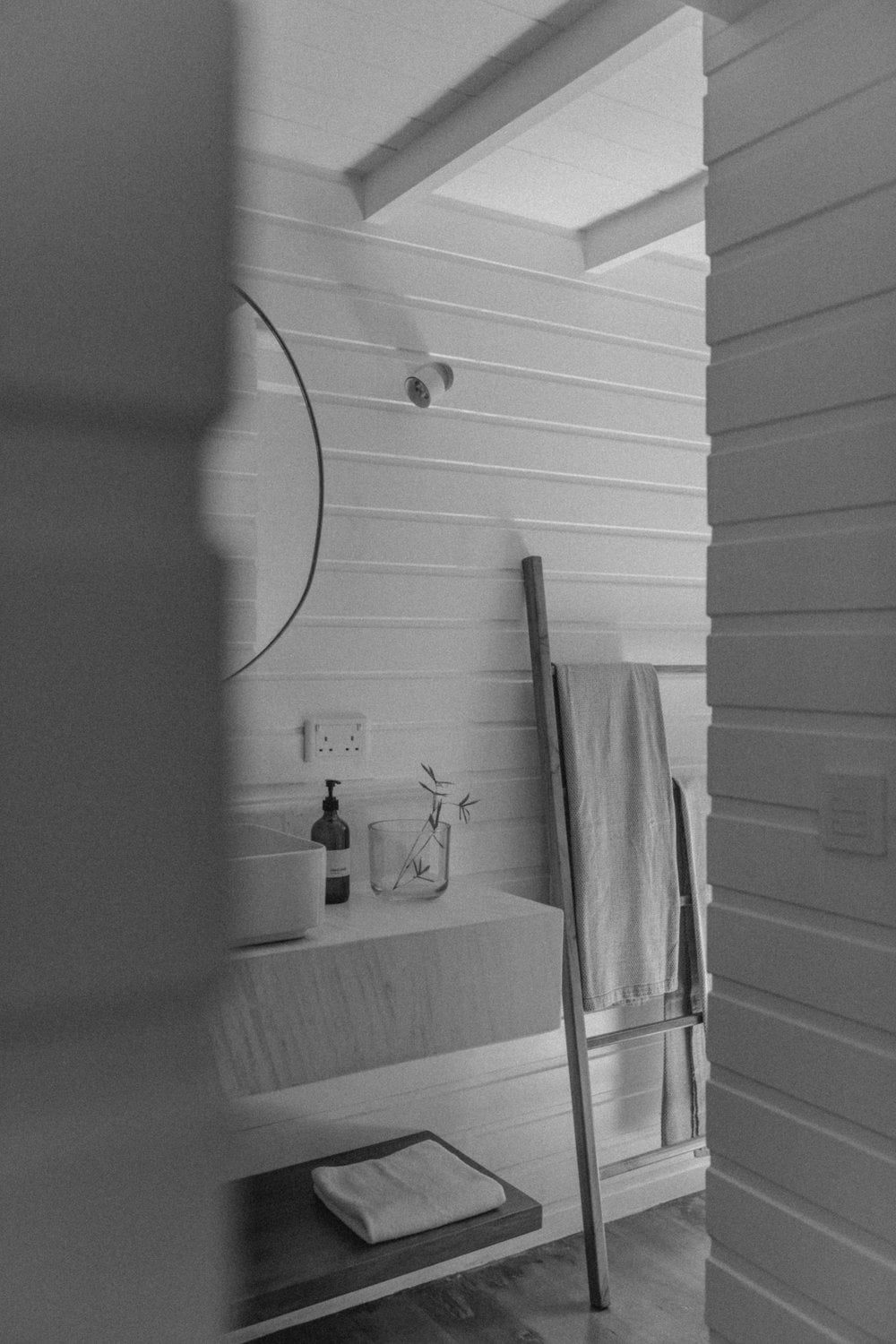 Kurulu Bay, Asia — Minimalist bathroom alcove with horizontal shiplap walls, skylight, floating shelf, dark towel rack, soap dispenser, and natural light casting clean shadows.