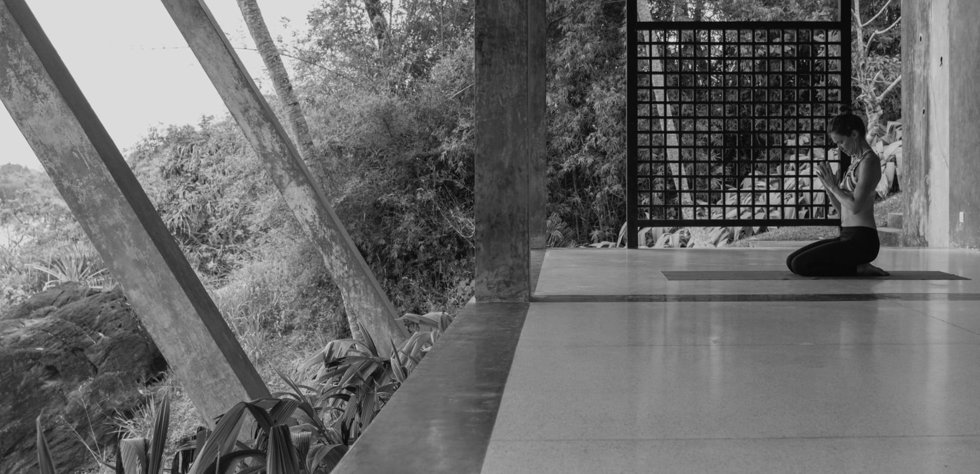 Yoga Retreats, Asia — Woman meditating in minimalist concrete pavilion overlooking dense tropical forest, lattice screen filtering natural light.