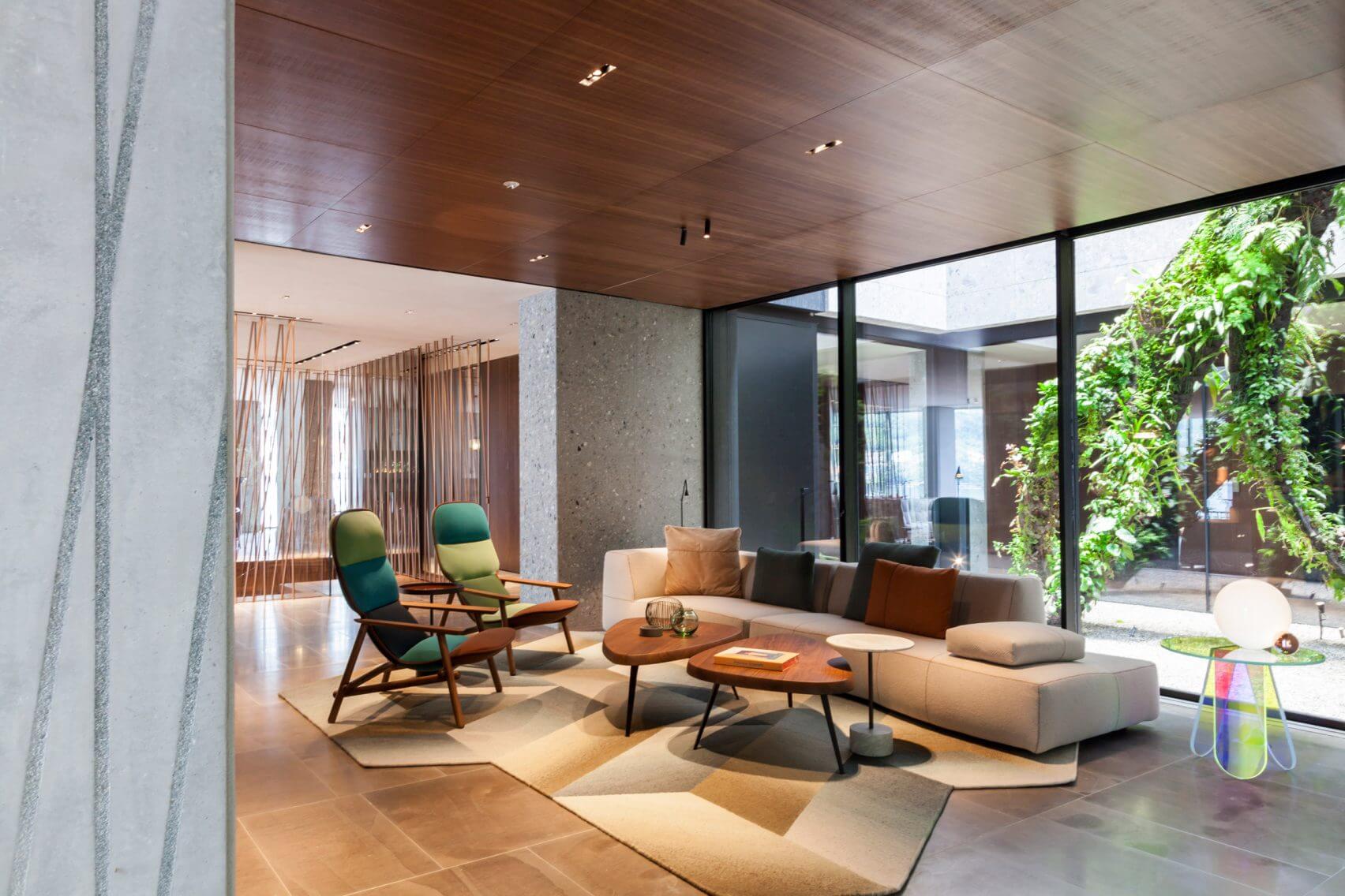 il Sereno Lago di Como, Europe — Modern living room with mid-century lounge chairs, cream sectional sofa, wooden coffee table, concrete walls, and floor-to-ceiling windows opening to landscaped courtyard.