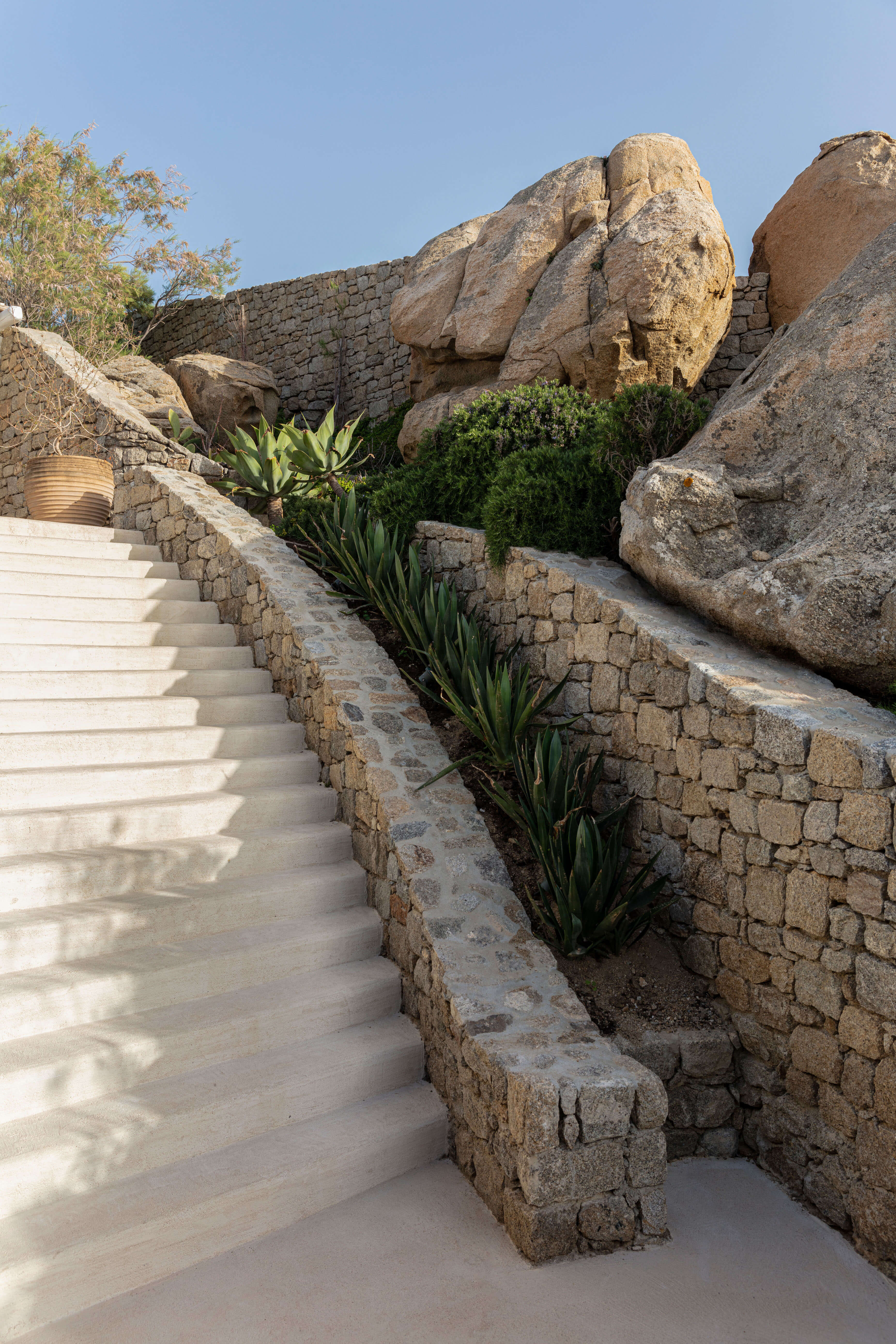 Villa Tama, Europe — Stone staircase ascending between granite boulders and dry-stacked walls with agave plants, clear sky, Mediterranean architecture integrated with natural rock formations.