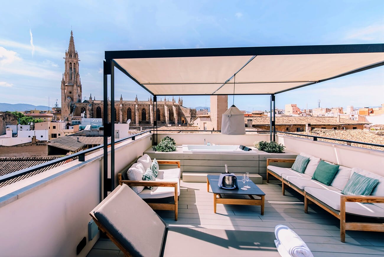 Sant Francesc Hotel Singular, Balearic Islands, Spain — Rooftop terrace with wooden loungers, retractable shade canopy, Gothic church spire, terracotta cityscape, afternoon light.