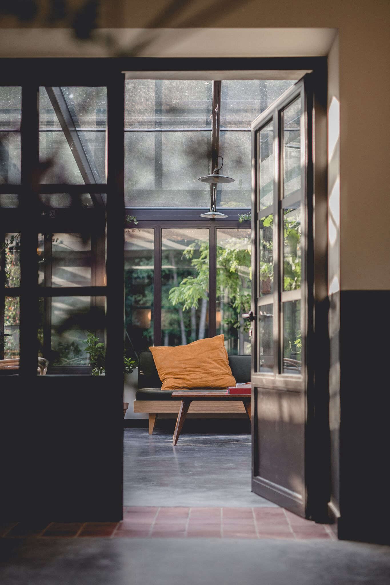 Le Barn, Europe — Black-framed glass doors opening to mid-century lounge chair with mustard cushion, garden views, natural light casting geometric shadows.