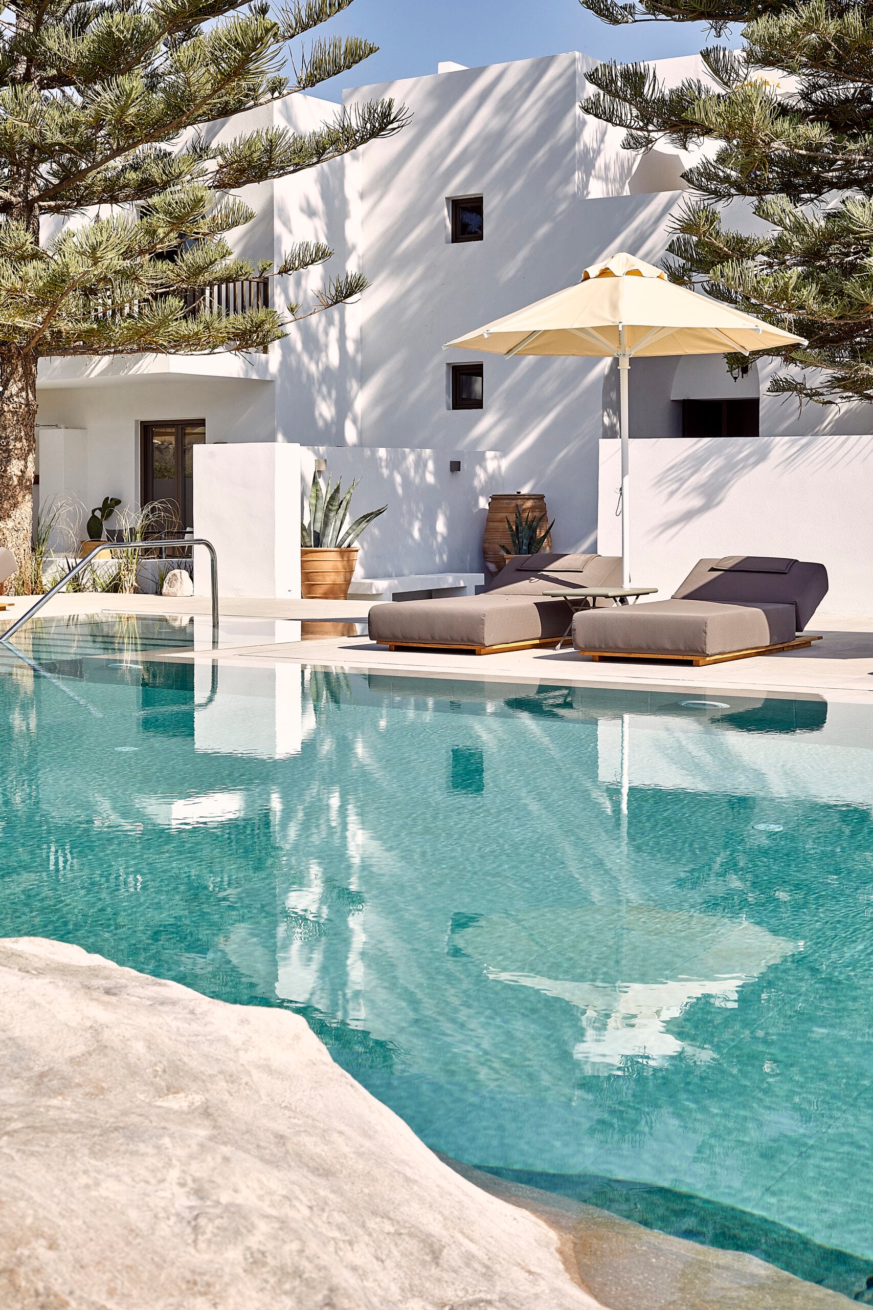 Parīlio Hotel, Europe — Whitewashed architecture with turquoise pool, sun loungers, cream umbrella, pine trees, and minimalist Mediterranean poolside design.