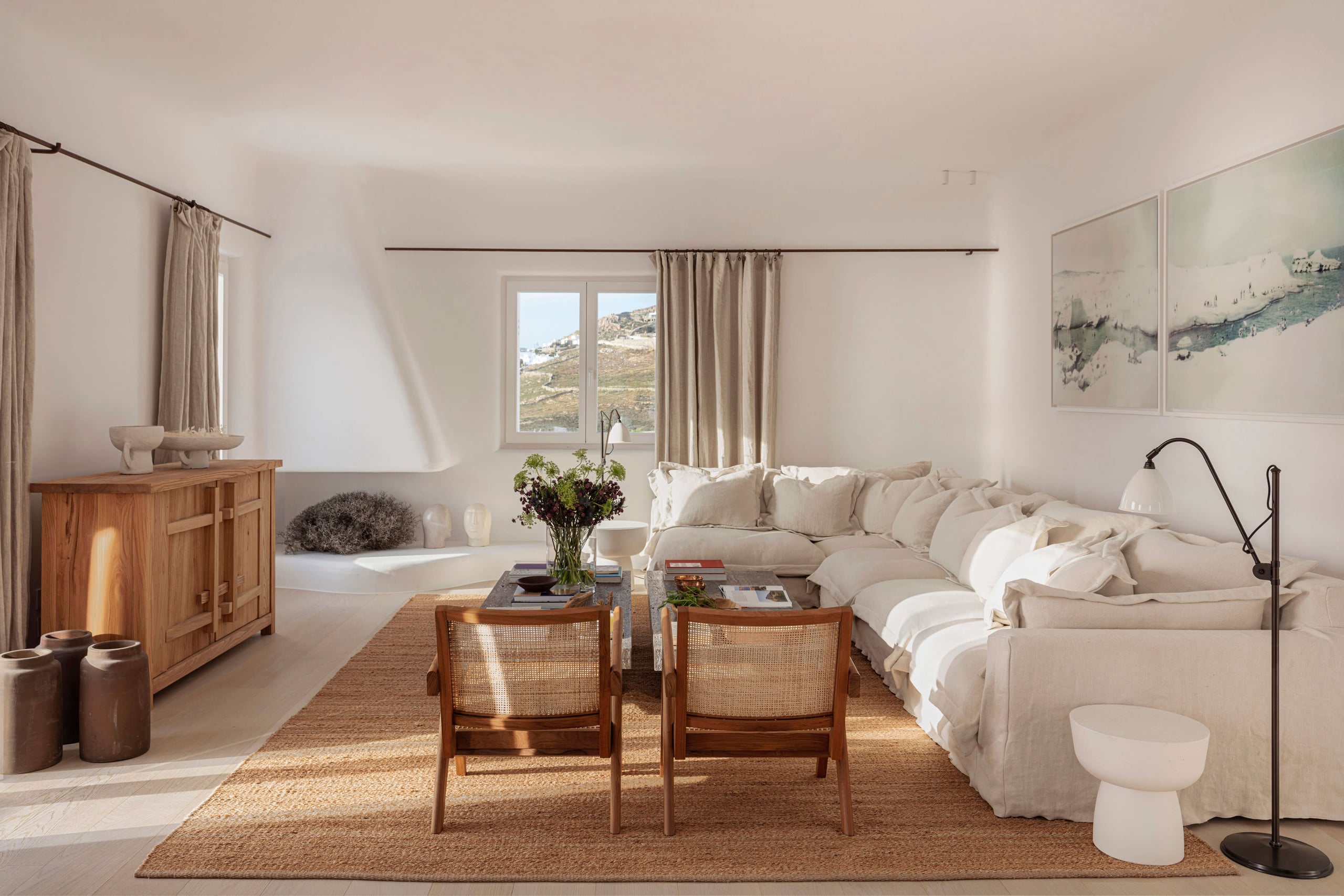 Villa Tama, Europe — Minimalist bedroom with cream linen sofa, woven chairs, wooden furniture, and Mediterranean landscape views through expansive windows.