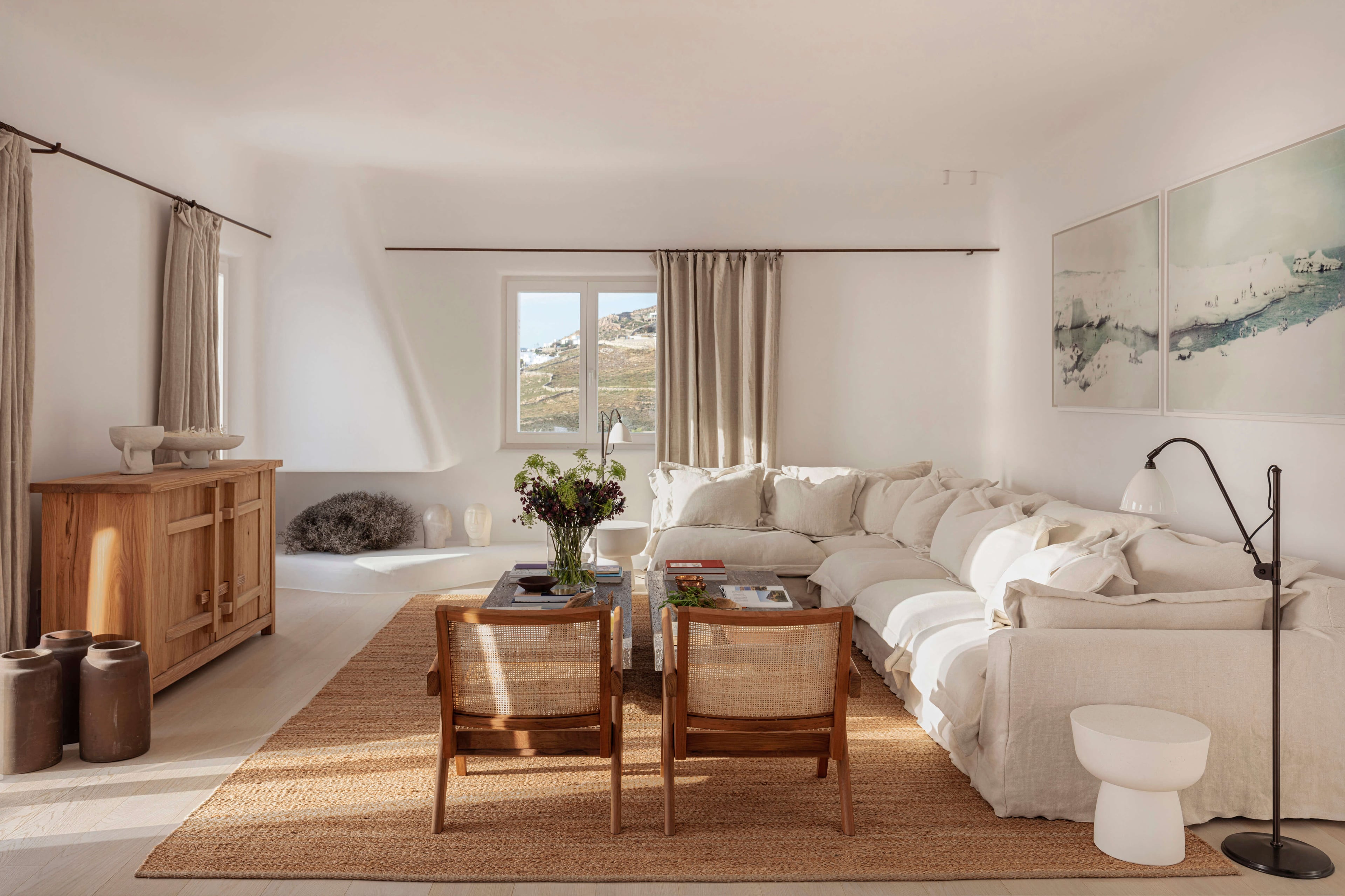 Villa Tama, Europe — Minimalist bedroom with cream linen sofa, woven chairs, wooden furniture, and Mediterranean landscape views through expansive windows.