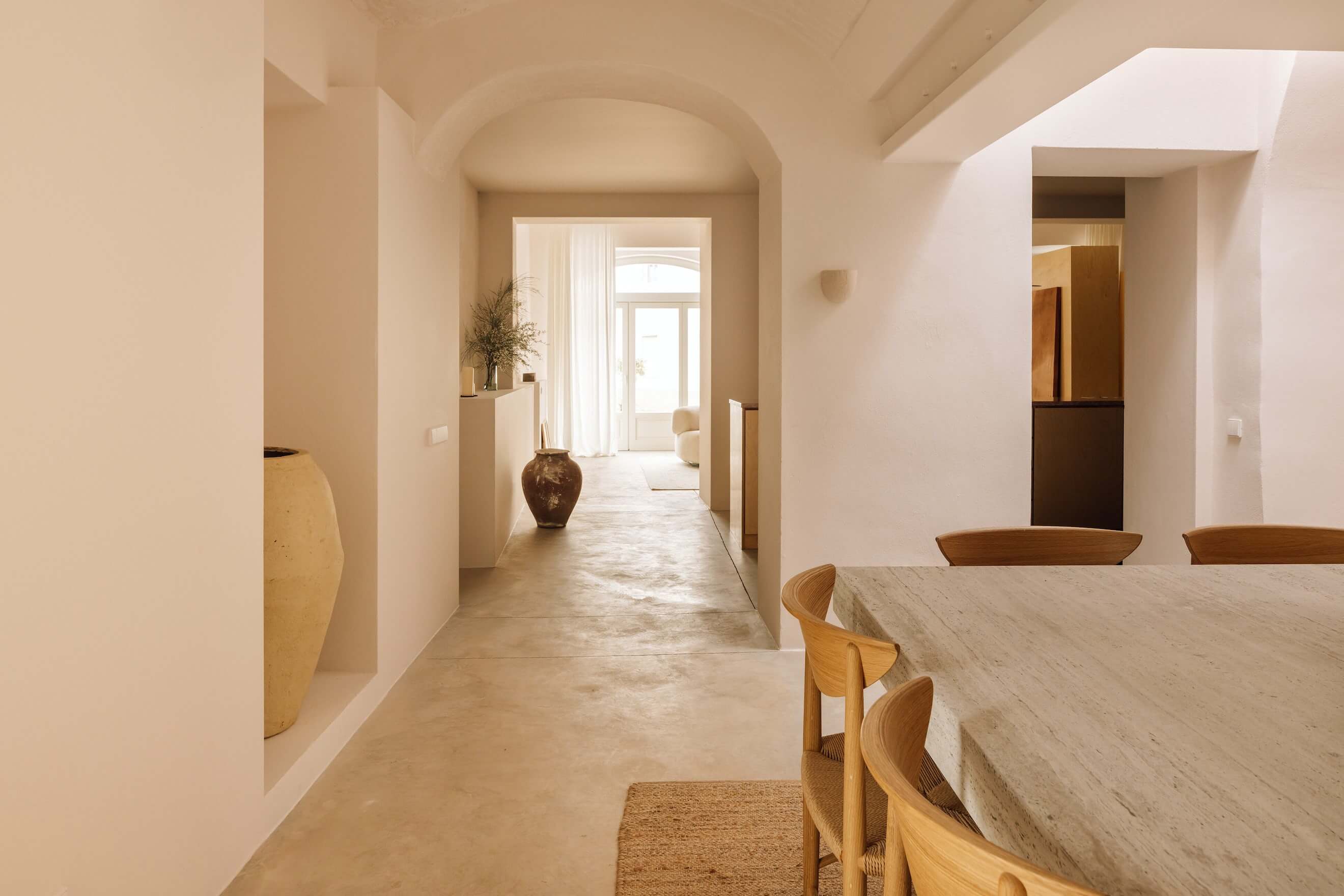 Casa Quatro, Europe — Minimalist hallway with arched ceiling, natural light, wooden chairs, terracotta vessels, limestone flooring, and warm neutral palette.