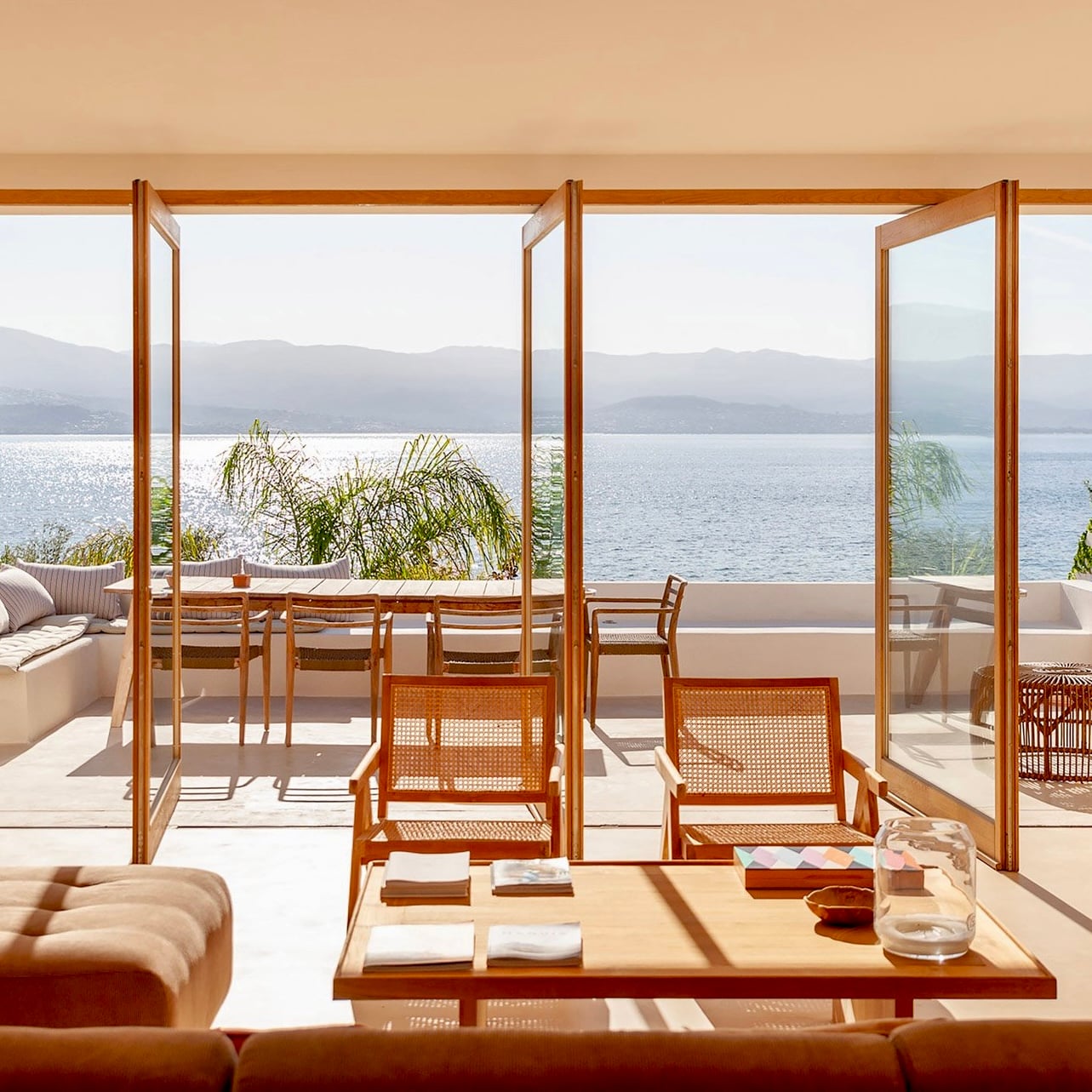 Casa Santa Teresa, Europe — Minimalist waterfront living room with wooden frames, rattan chairs, and panoramic lake views across mountains in soft natural light.