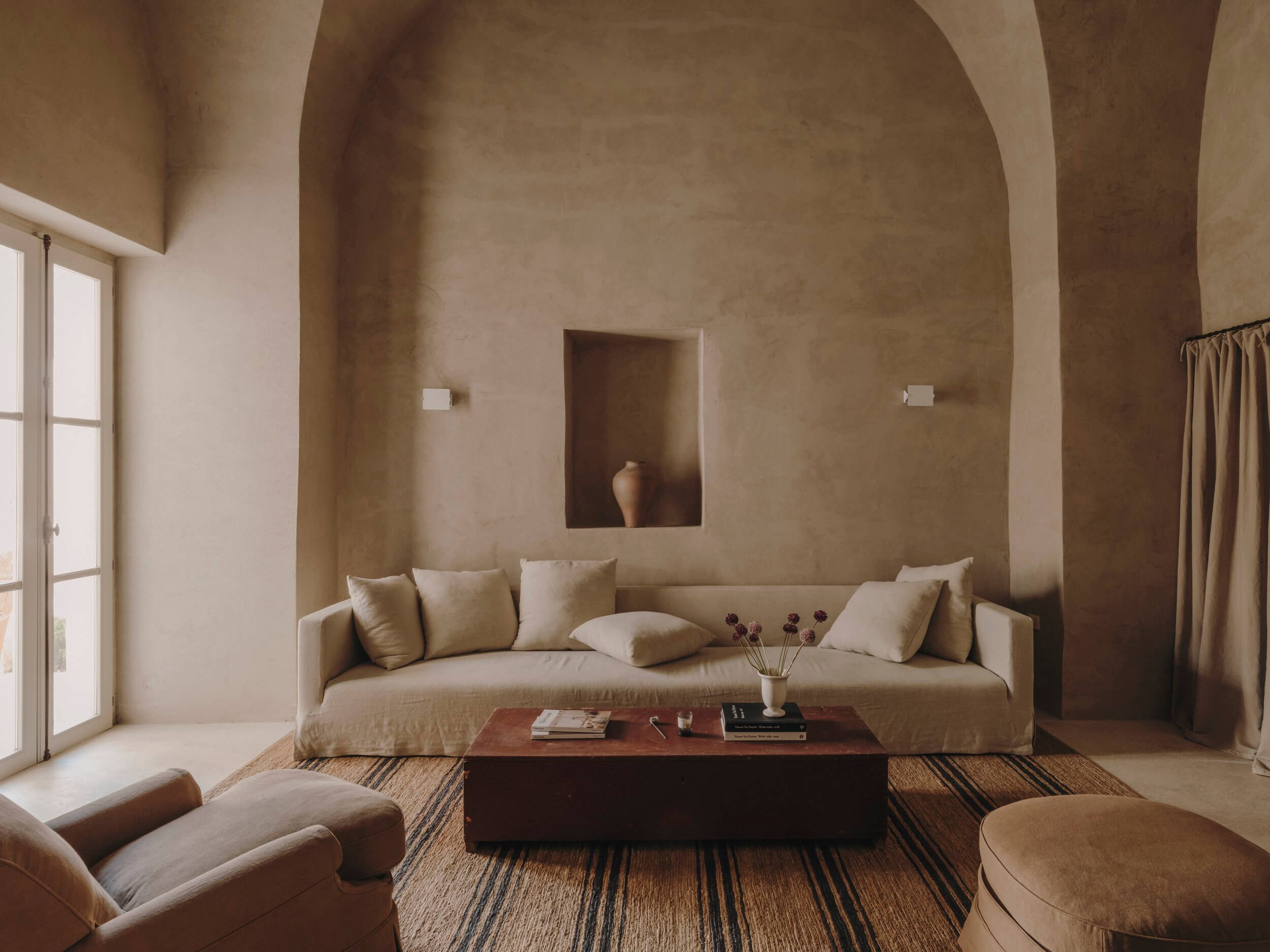 Casa Soleto, Europe — Minimalist living room with arched plaster walls, cream linen sofa, terracotta accents, and natural light from floor-to-ceiling doors.