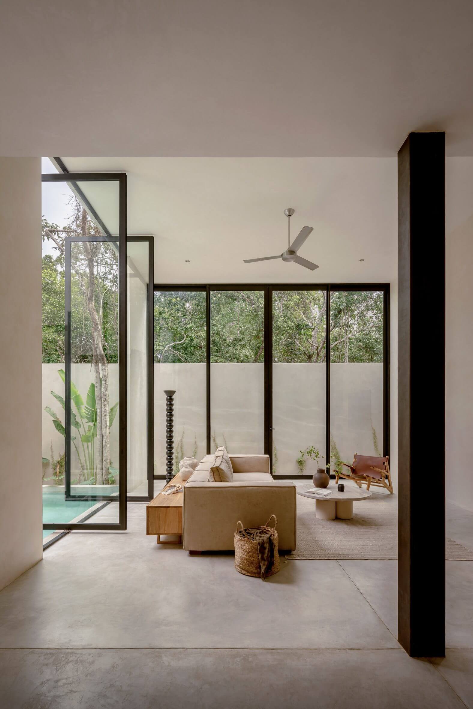 Villa Cava, Mexico — Minimalist living room with black-framed glass doors, natural light, ceiling fan, and views of tropical forest through floor-to-ceiling windows.