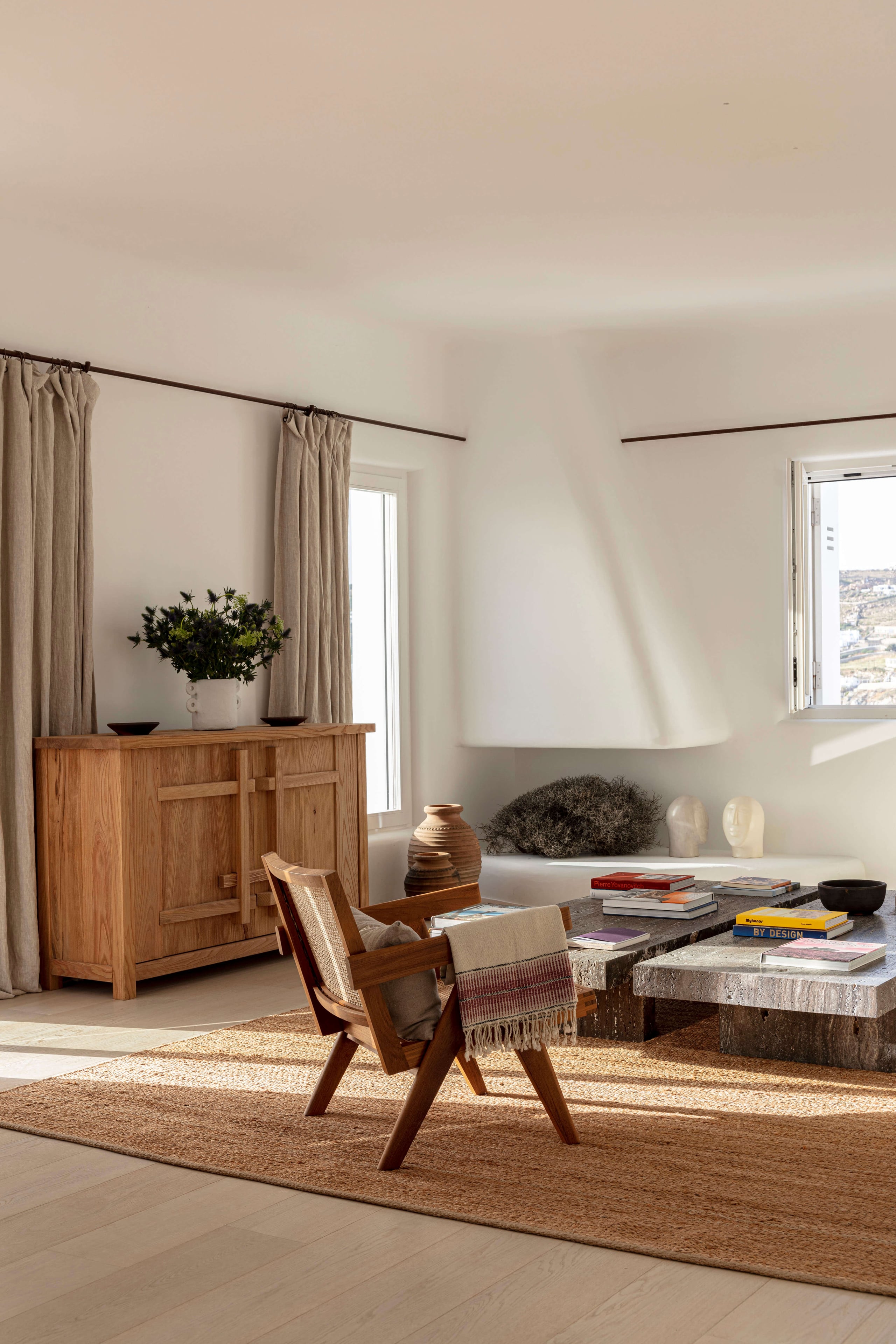Villa Tama, Europe — Minimalist living room with wooden mid-century chair, stone coffee table, linen curtains, natural light, neutral palette.