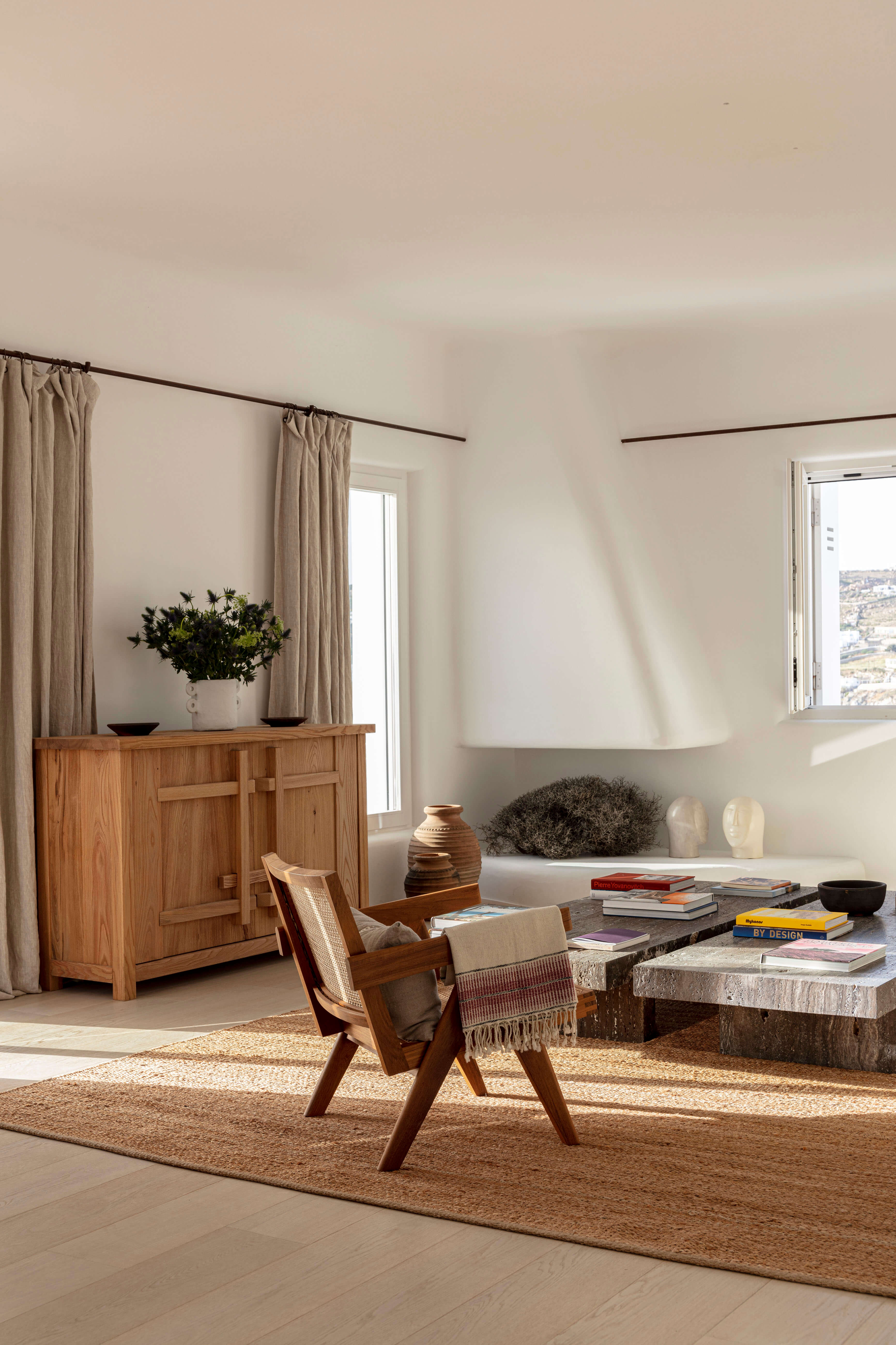 Villa Tama, Europe — Minimalist living room with wooden mid-century chair, stone coffee table, linen curtains, natural light, neutral palette.