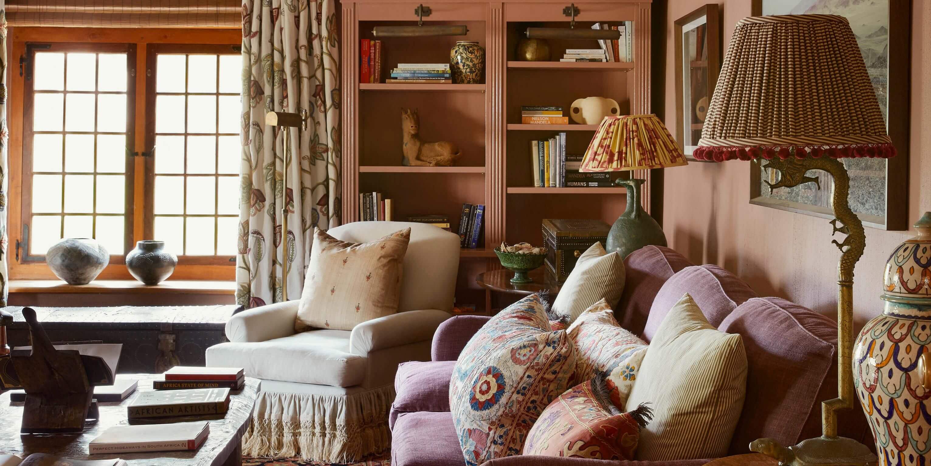 Sterrekopje, Africa — Warm library lounge with terracotta shelving, cream armchair, plush mauve seating, patterned pillows, brass floor lamp, and natural light from wood-framed windows.