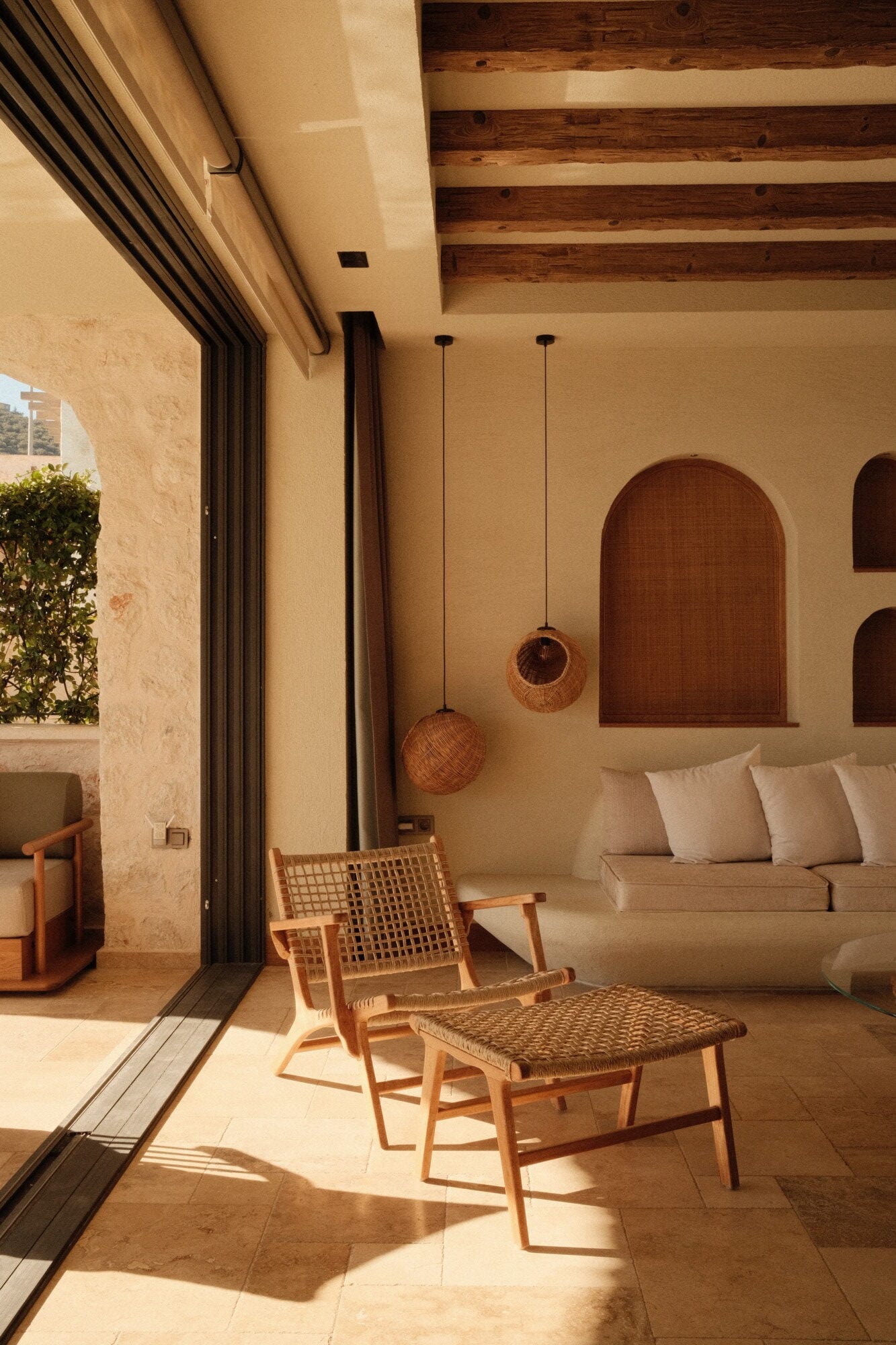 Villa Thalia by Muses Kalkan, Kalkan, Turkey — Minimalist terrace with woven pendant lights, wooden lounge chair, cream sofa, arched niches, and natural stone walls opening to Mediterranean garden.
