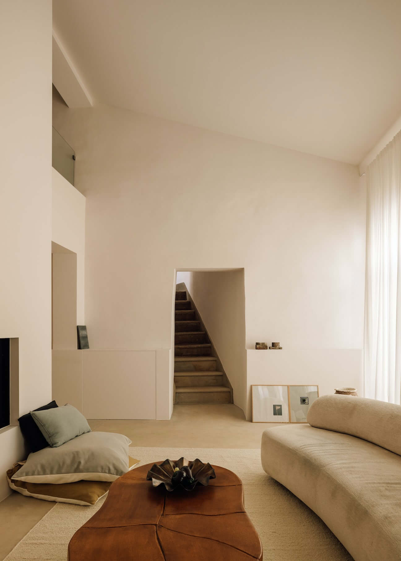 Casa Quatro, Europe — Minimalist living space with cream walls, sculptural seating, cognac leather poufs, and staircase opening to upper level, flooded with natural light.