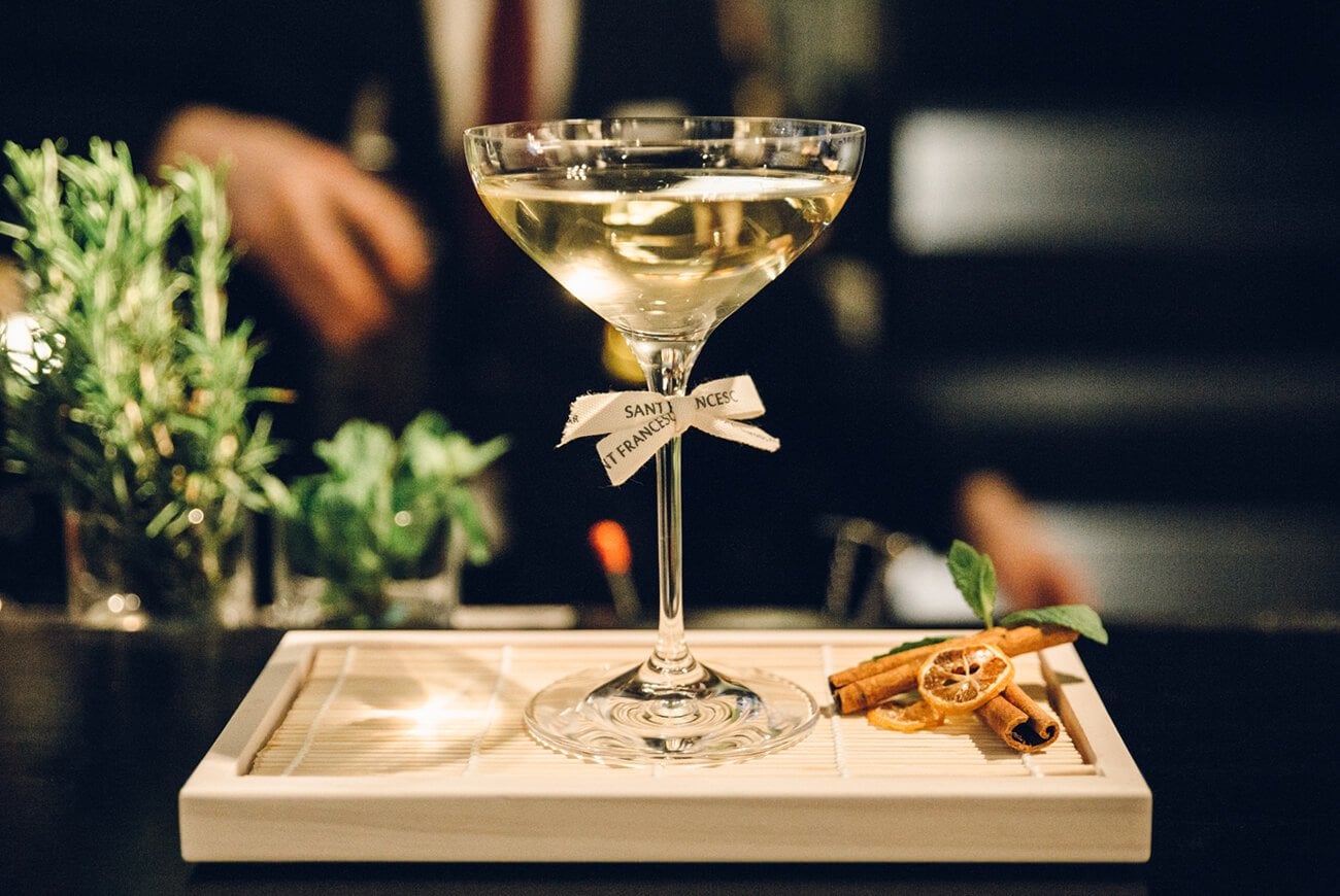 Sant Francesc Hotel Singular, Balearic Islands, Spain — pale yellow cocktail with herb garnish and ribbon label on white tray, candied citrus and cinnamon beside, dark ambient bar setting.