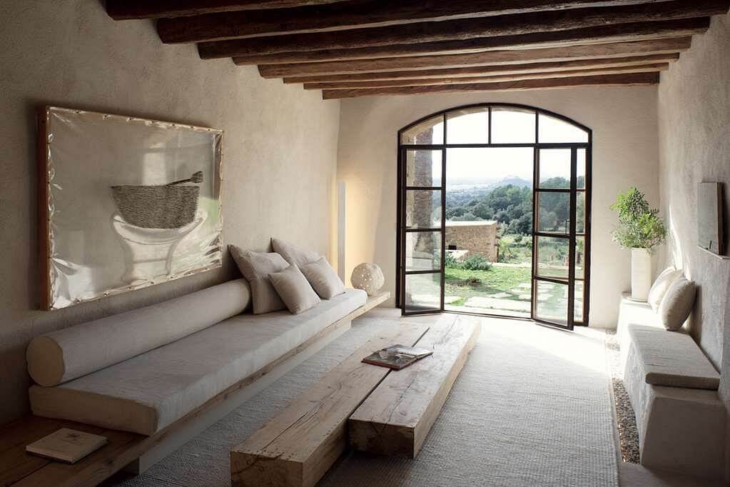 Es Racó d'Artà, Balearic Islands, Spain — Minimalist bedroom with exposed timber beams, arched glass doors opening to landscape, linen daybed, whitewashed walls, natural light.