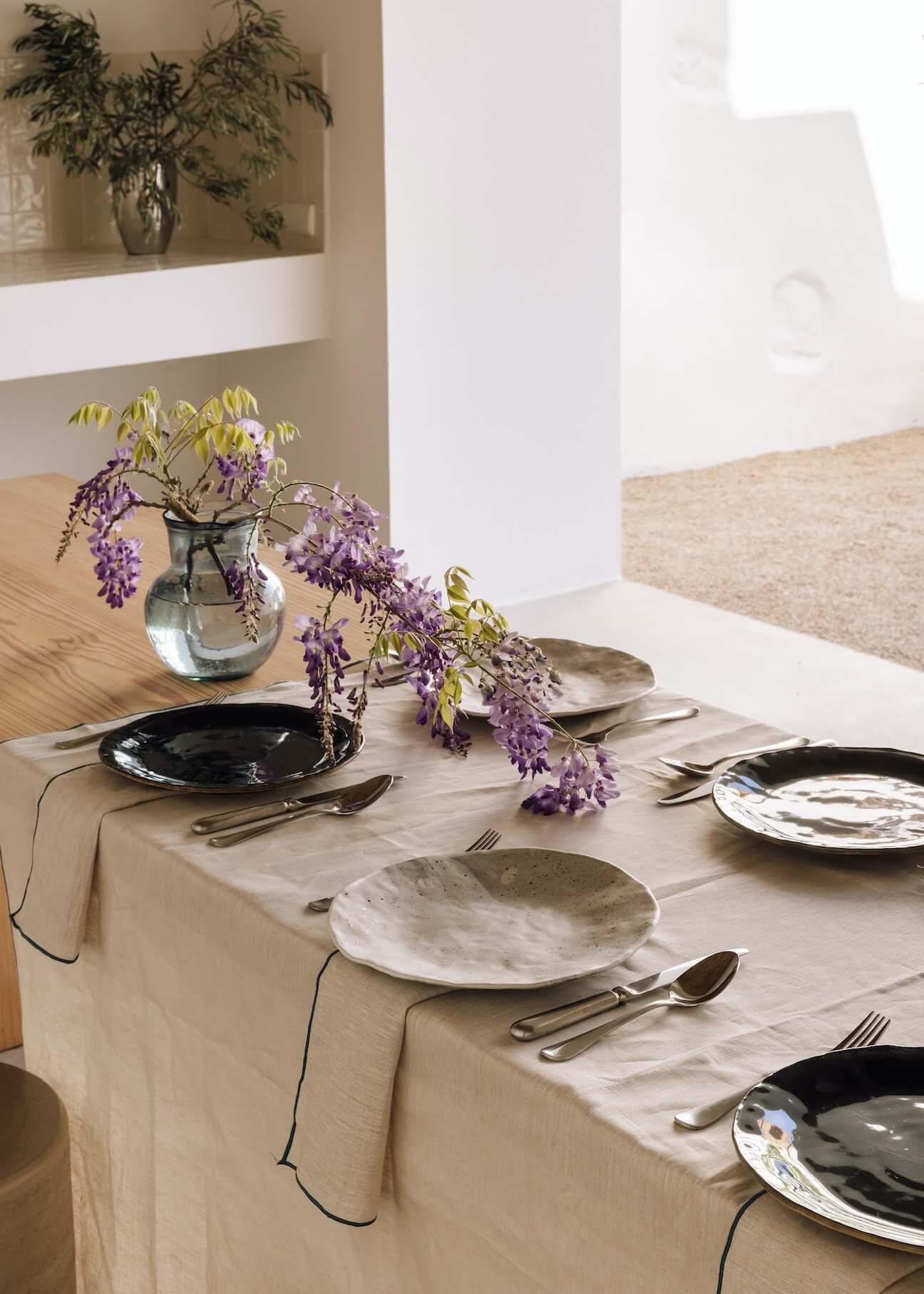 Casa Quatro, Europe — Minimalist dining table with woven linen runner, purple wisteria arrangement, and mixed ceramic and black dinnerware in bright natural light.