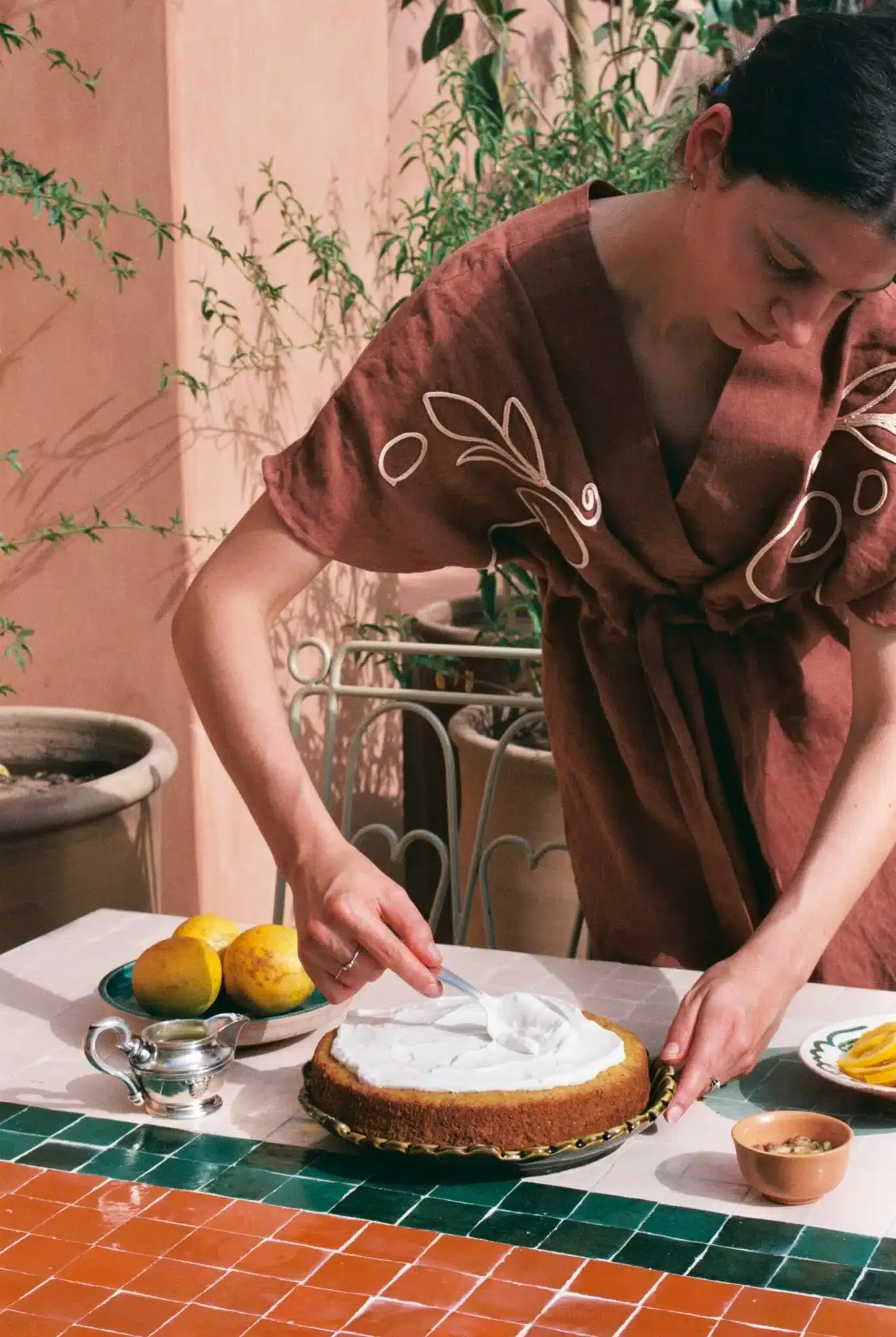 Rosemary, Africa — Woman in terracotta linen garnishing citrus cake with cream at tiled table, green climbing vines and pink stucco walls behind.